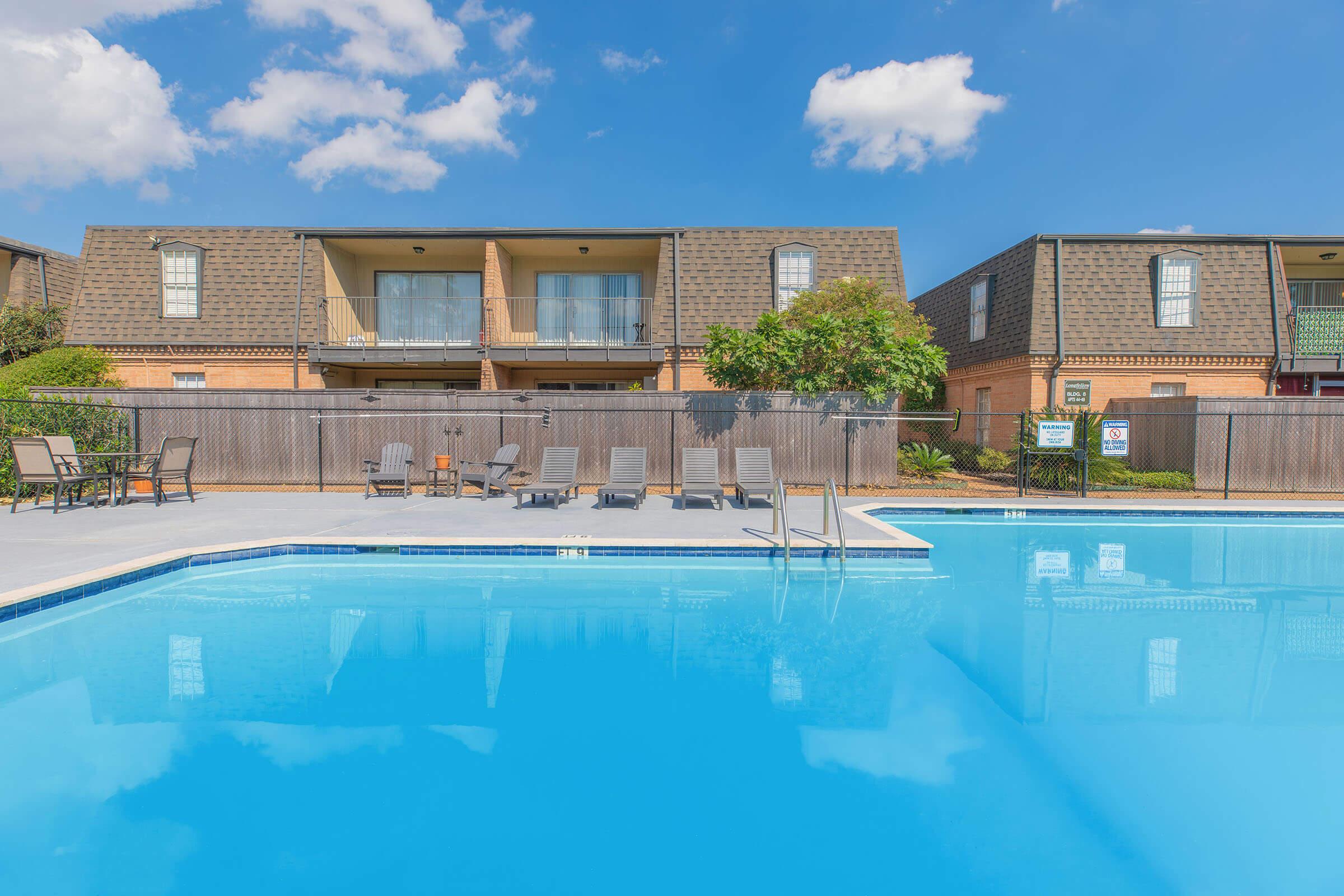 Swimming pool with clear blue water surrounded by lounge chairs. In the background, two-story apartment buildings feature balconies, set against a bright blue sky with a few clouds. A wooden fence encloses the pool area, adding to the outdoor relaxation space.
