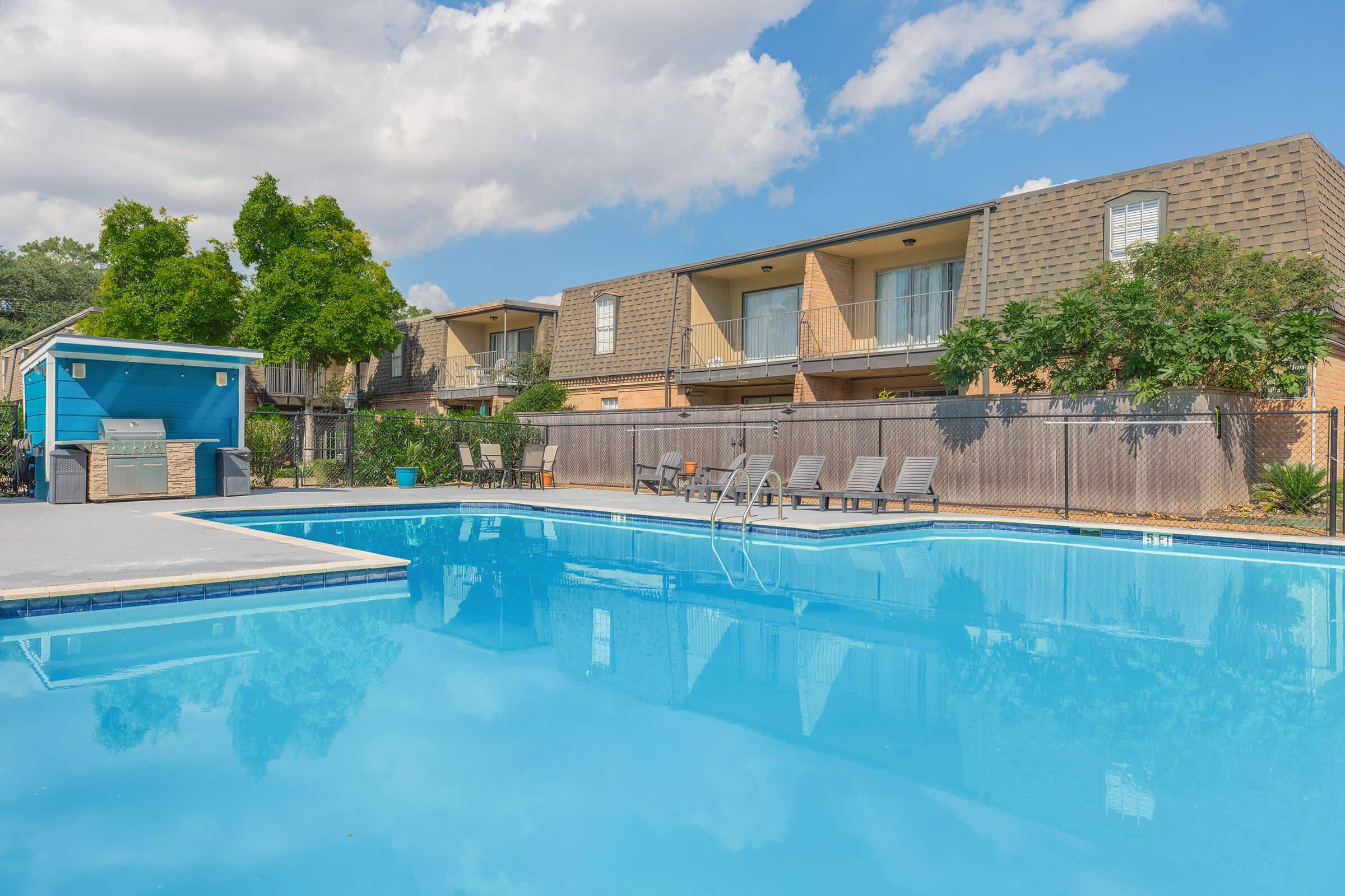 A clear blue swimming pool surrounded by lounge chairs, with a fence and greenery. In the background, there are apartment buildings with balconies and a small outdoor kitchen area. The sky is partly cloudy, adding a vibrant feel to the scene.