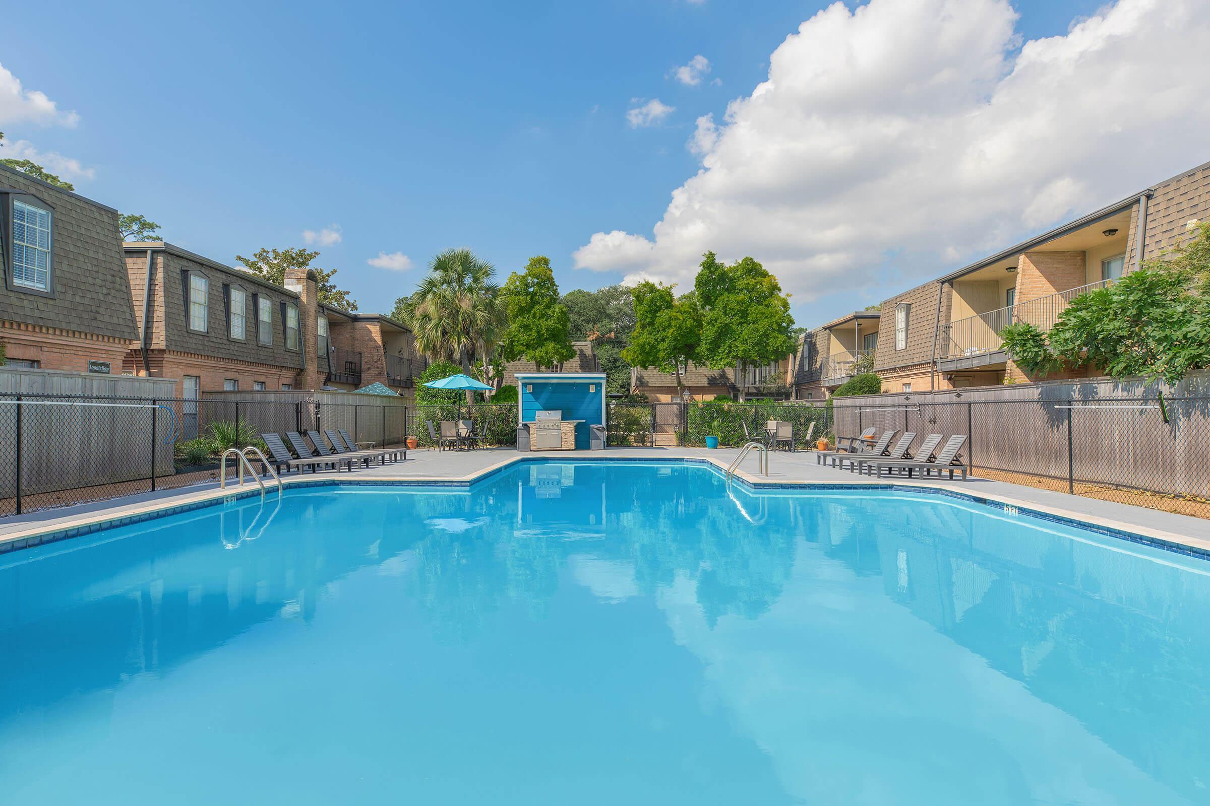 A clear blue swimming pool surrounded by lounge chairs, with palm trees and green umbrellas in the background. Residential buildings framed by lush landscaping are visible behind the pool, under a bright blue sky with fluffy white clouds.