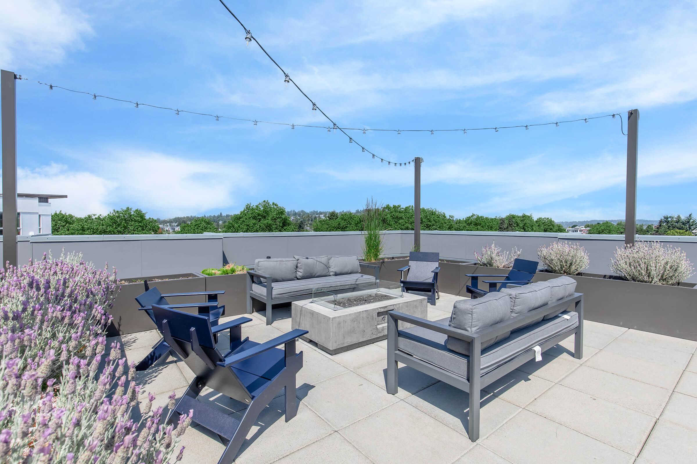 A spacious rooftop terrace featuring comfortable seating arrangements with gray cushions and navy blue chairs. In the center, there is a modern fire pit surrounded by greenery and decorative plants. The terrace is illuminated by string lights against a clear blue sky, providing a cozy outdoor ambiance.