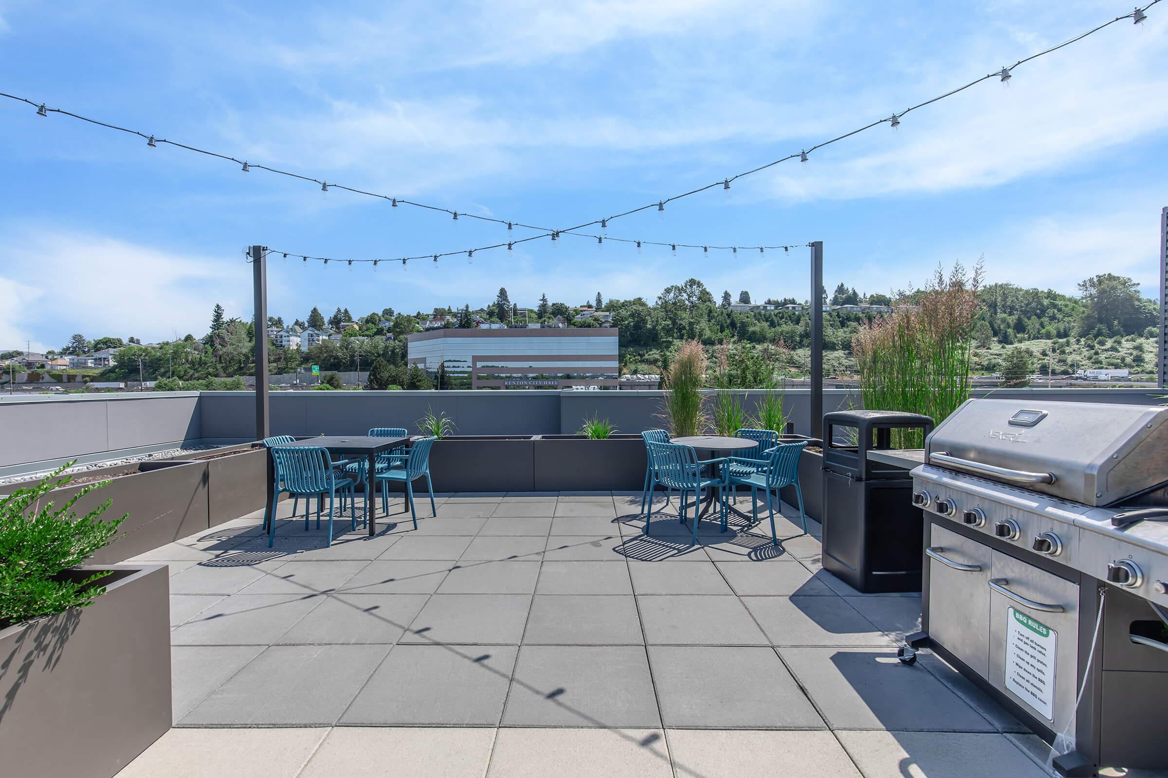 A rooftop patio featuring a barbecue grill, outdoor dining tables with chairs, and decorative plants. String lights hang overhead, adding ambiance to the space, with a scenic view of trees and buildings in the background under a bright blue sky.