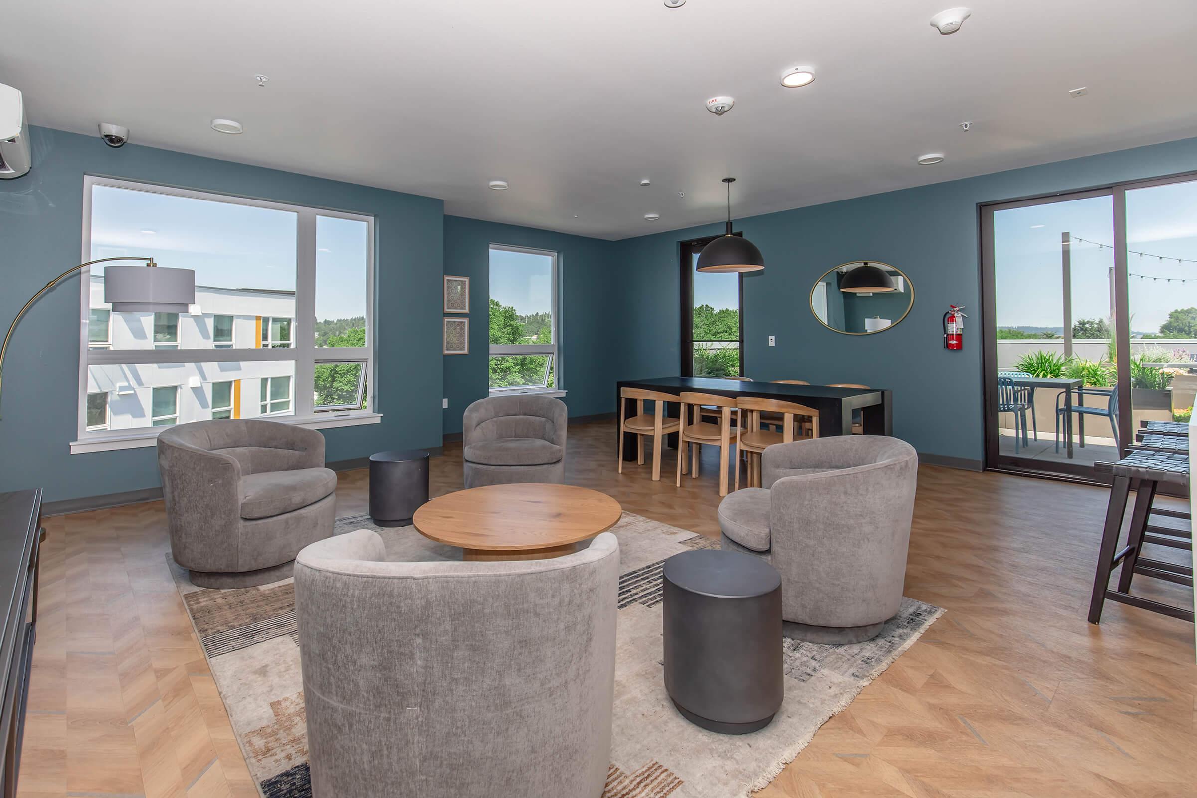A modern lounge area featuring four plush gray armchairs arranged around a wooden coffee table. A dining table with chairs is positioned nearby. The walls are painted in a soft blue color. Large windows offer views of greenery outside, and there are warm wooden floors throughout the space.