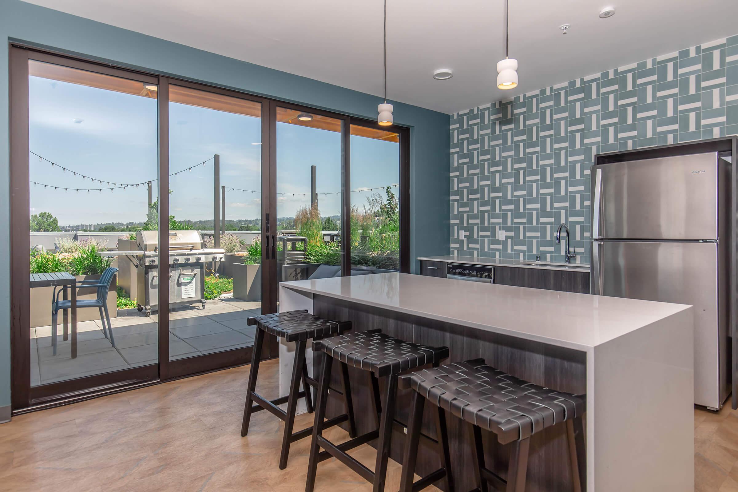 Modern kitchen featuring a large island with bar stools, sleek stainless steel appliances, and a stylish patterned backsplash. Large sliding glass doors open to an outdoor patio with greenery and a grill, providing a bright and spacious atmosphere.