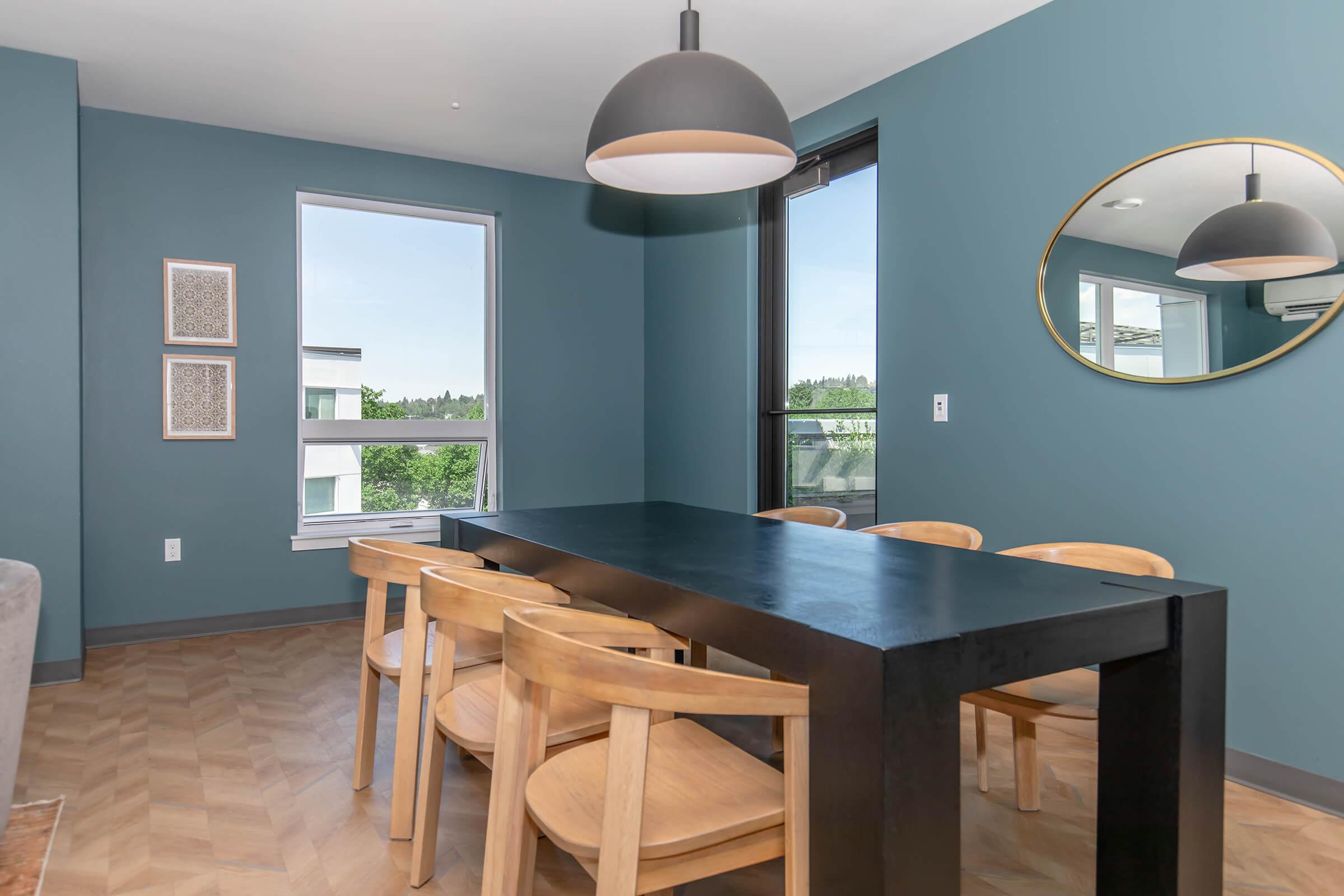 Modern dining room featuring a sleek black table surrounded by wooden chairs. The walls are painted a cool blue, and there are large windows allowing natural light. A round mirror and two framed artworks add decorative touches, while greenery is visible outside through the windows.