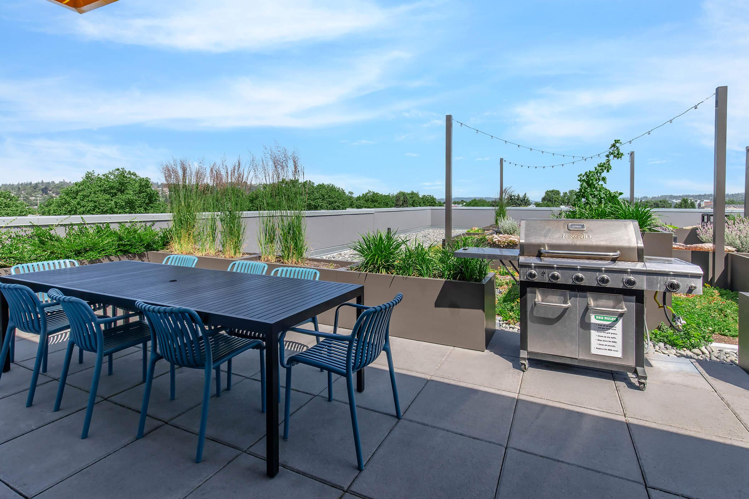 A spacious outdoor terrace featuring a large dining table with blue chairs, a stainless steel grill, and lush greenery in planters. The setting is bright and airy, with a clear blue sky and a landscaped view, perfect for gatherings and barbecues.