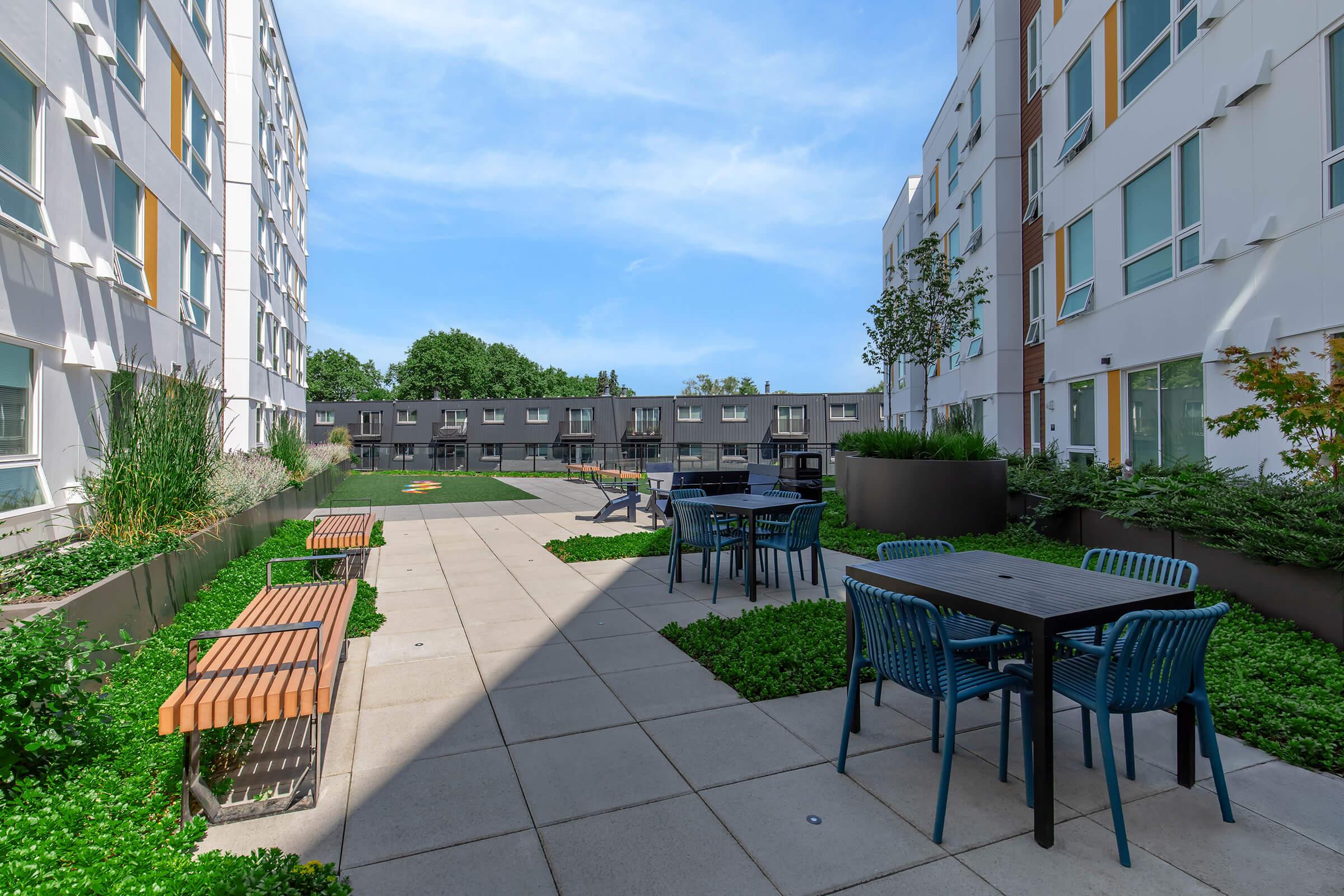 A modern outdoor courtyard with green landscaping. Features include benches, tables with chairs, and a lawn area, surrounded by contemporary apartment buildings. The sky is clear, providing a bright and inviting atmosphere.
