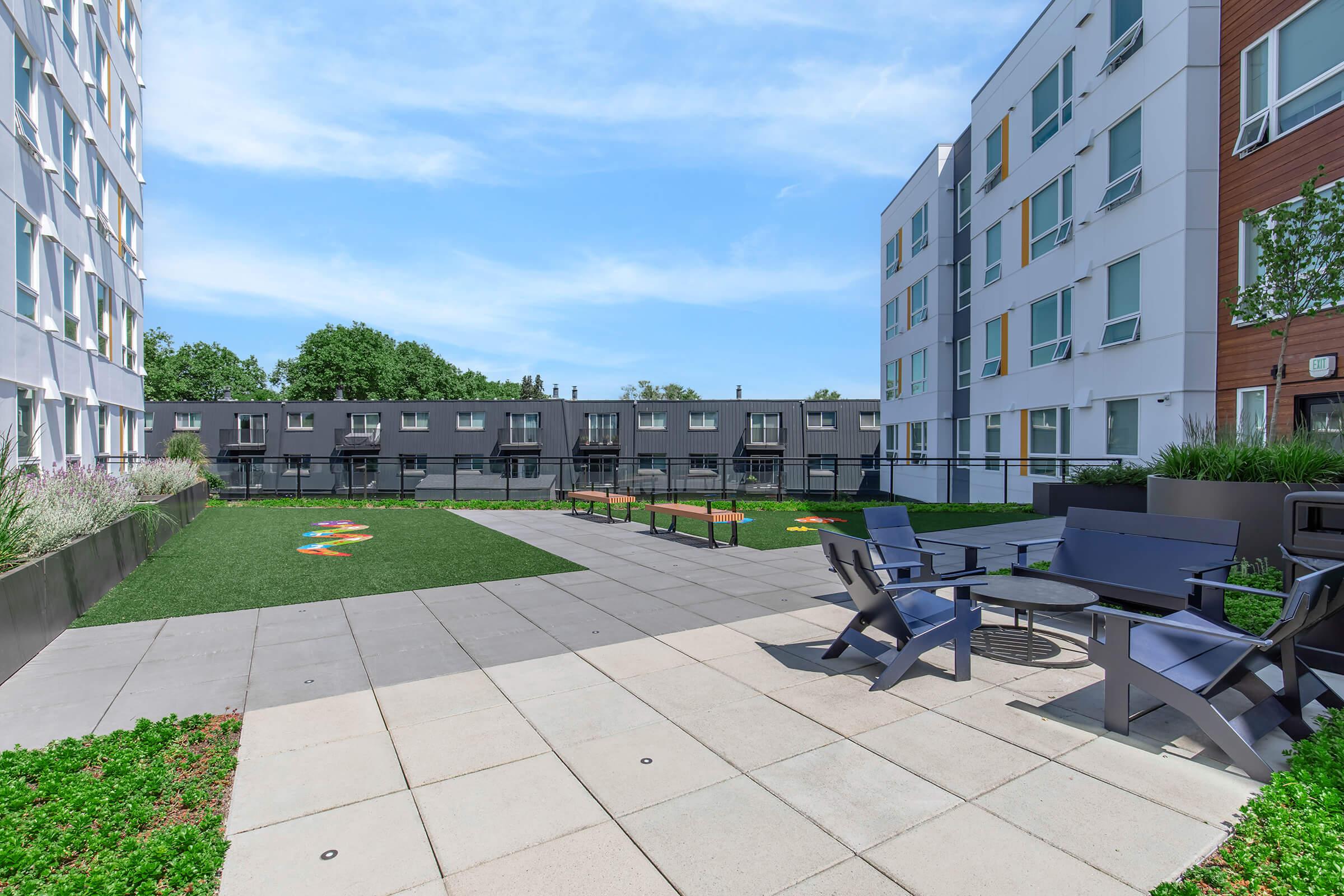 A well-maintained outdoor courtyard featuring a grassy area, modern benches, and seating arrangements. The space is surrounded by contemporary apartment buildings, with clear blue skies above, creating a welcoming and vibrant atmosphere for relaxation and socializing.