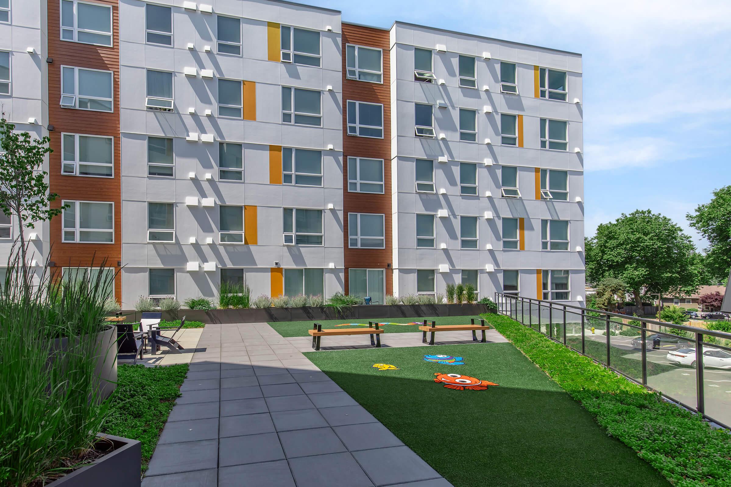 Modern residential building with multiple levels, featuring a landscaped outdoor area. The foreground includes a small play area with colorful designs on artificial grass, and benches for seating. The facade of the building is a mix of gray and wood accents, with large windows. Green plants and trees are visible around the space.