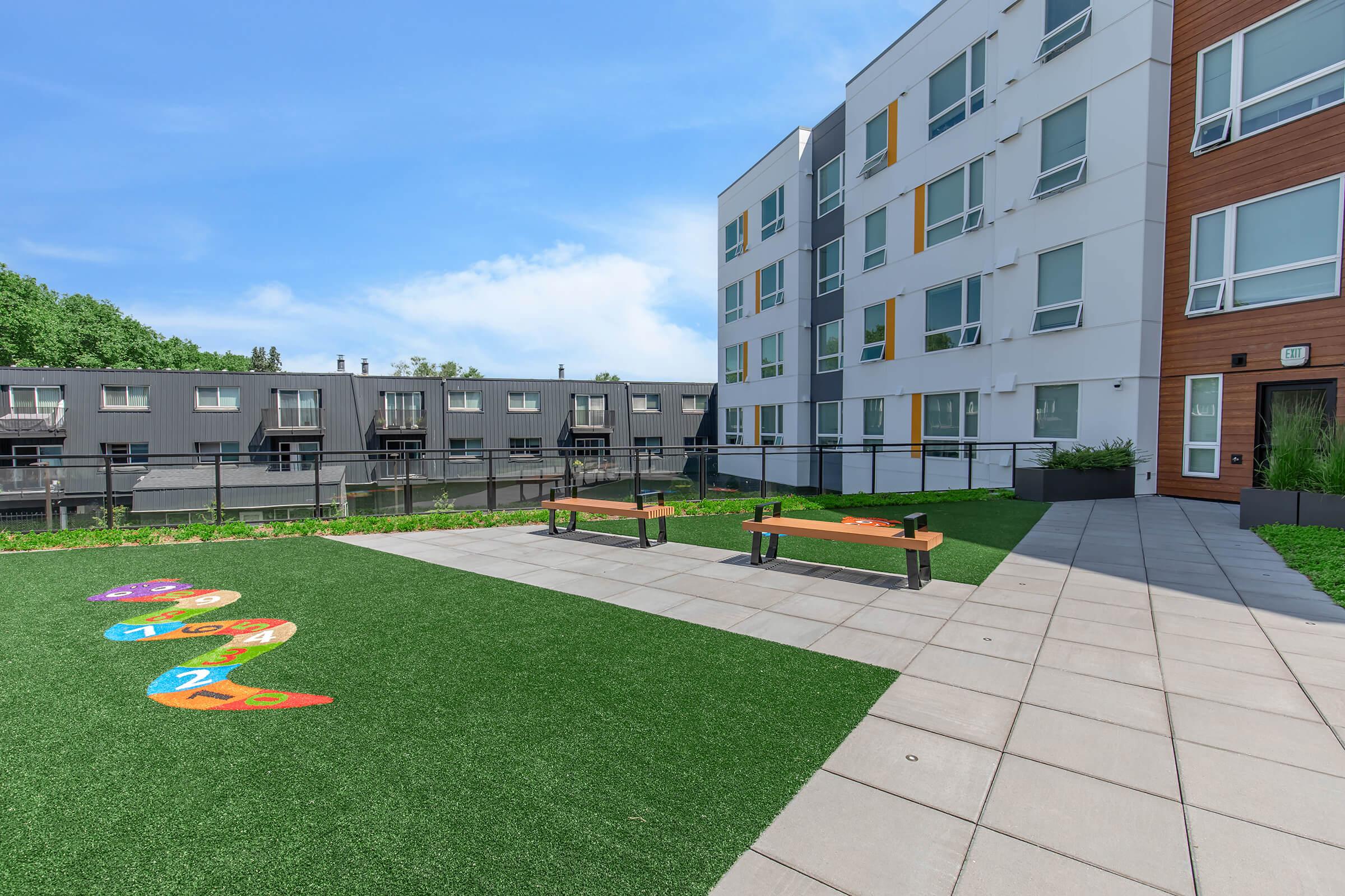 A modern outdoor courtyard with artificial grass, featuring colorful painted letters on the ground. There are two wooden benches positioned on a paved area, surrounded by a contemporary building and greenery in the background under a clear blue sky.