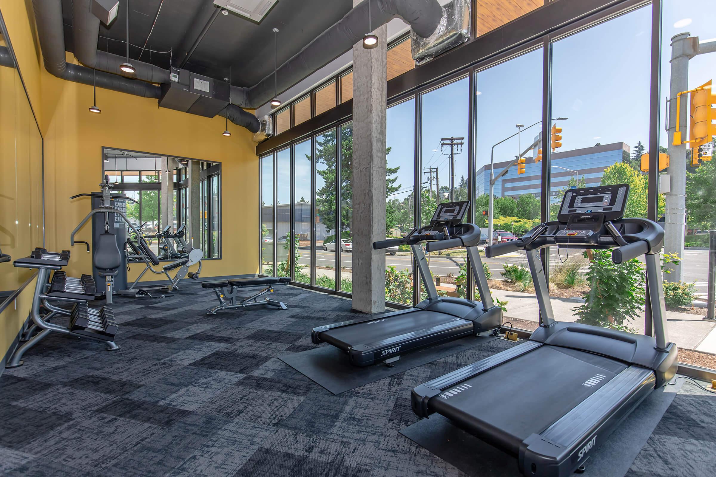 A modern gym interior featuring several treadmills and workout equipment. Large windows provide natural light and a view of the outdoor landscape. The walls are painted yellow, and there are black carpets on the floor.
