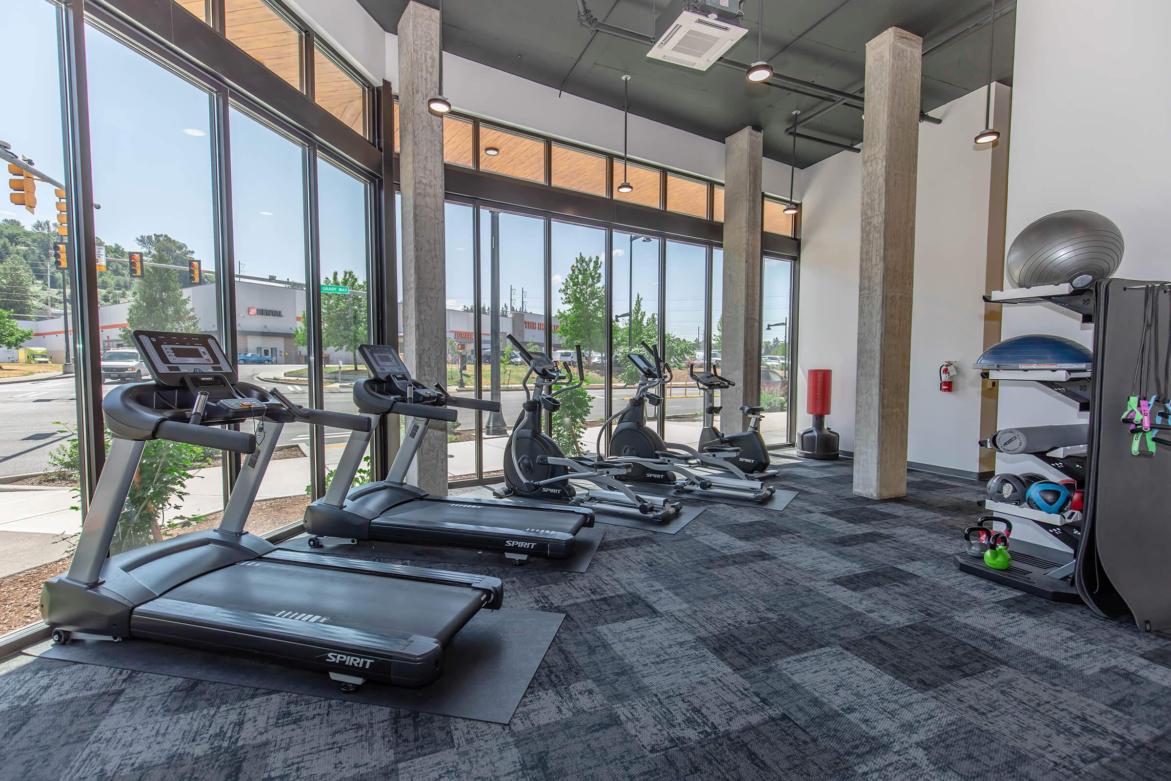 A modern gym interior featuring multiple treadmills and ellipticals lined up against large windows, allowing natural light to fill the space. The flooring is dark with a textured pattern, and there are various fitness accessories neatly organized on a storage unit against the wall.
