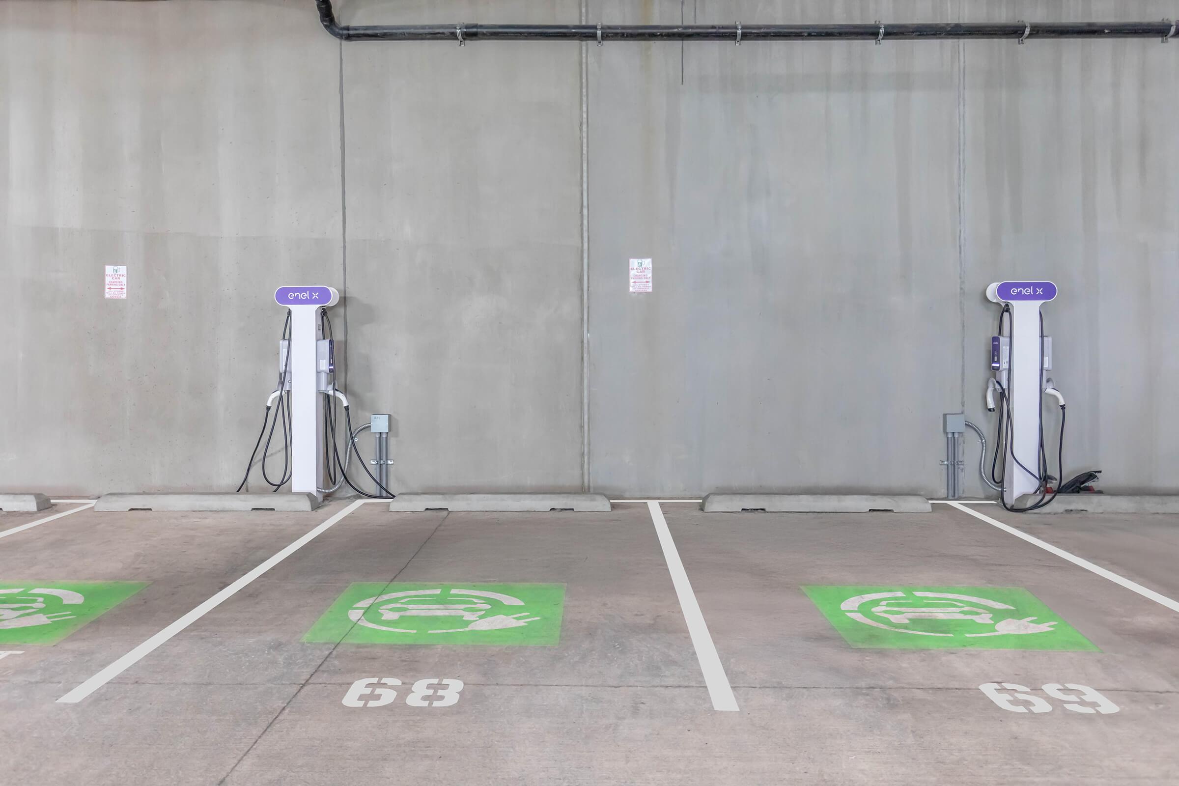 Two electric vehicle charging stations in a parking garage, marked with green symbols indicating charging spots. The stations are mounted on a concrete wall, with visible power cords hanging down. There are also parking spot numbers (63 and 64) visible on the ground.