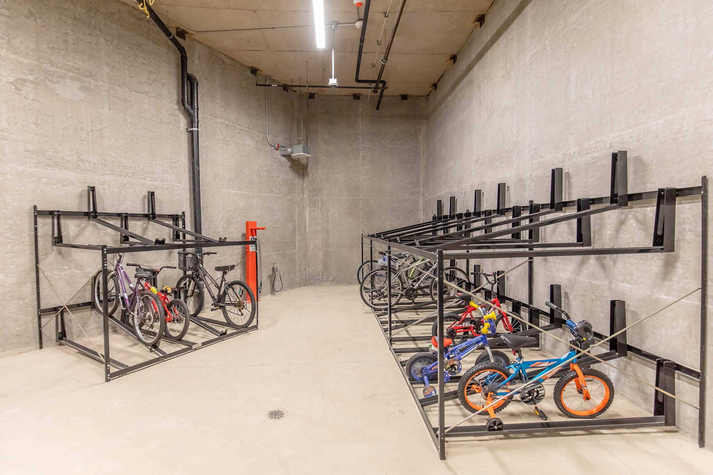 Clean and organized bike storage area with metal racks for bicycles. The space features concrete walls and flooring, with several bikes secured on the racks, including a mix of adult and children's bicycles in various colors. Bright overhead lighting illuminates the area.