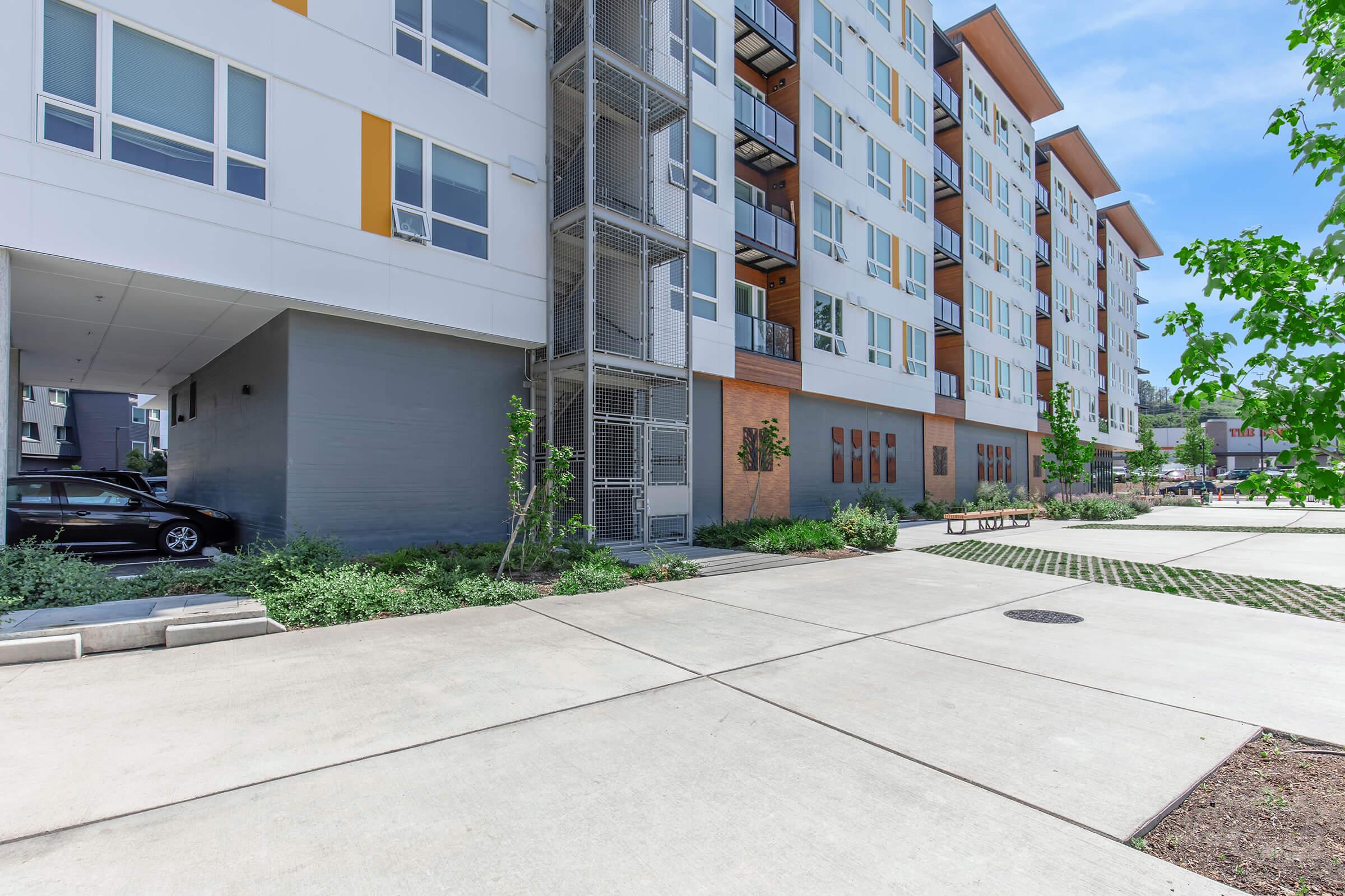 A modern apartment building with a light-colored exterior and large windows. The structure has multiple balconies and a landscaped area with greenery. A paved pathway leads to the entrance, and there is a parked car nearby. Bright blue skies are visible above the building.