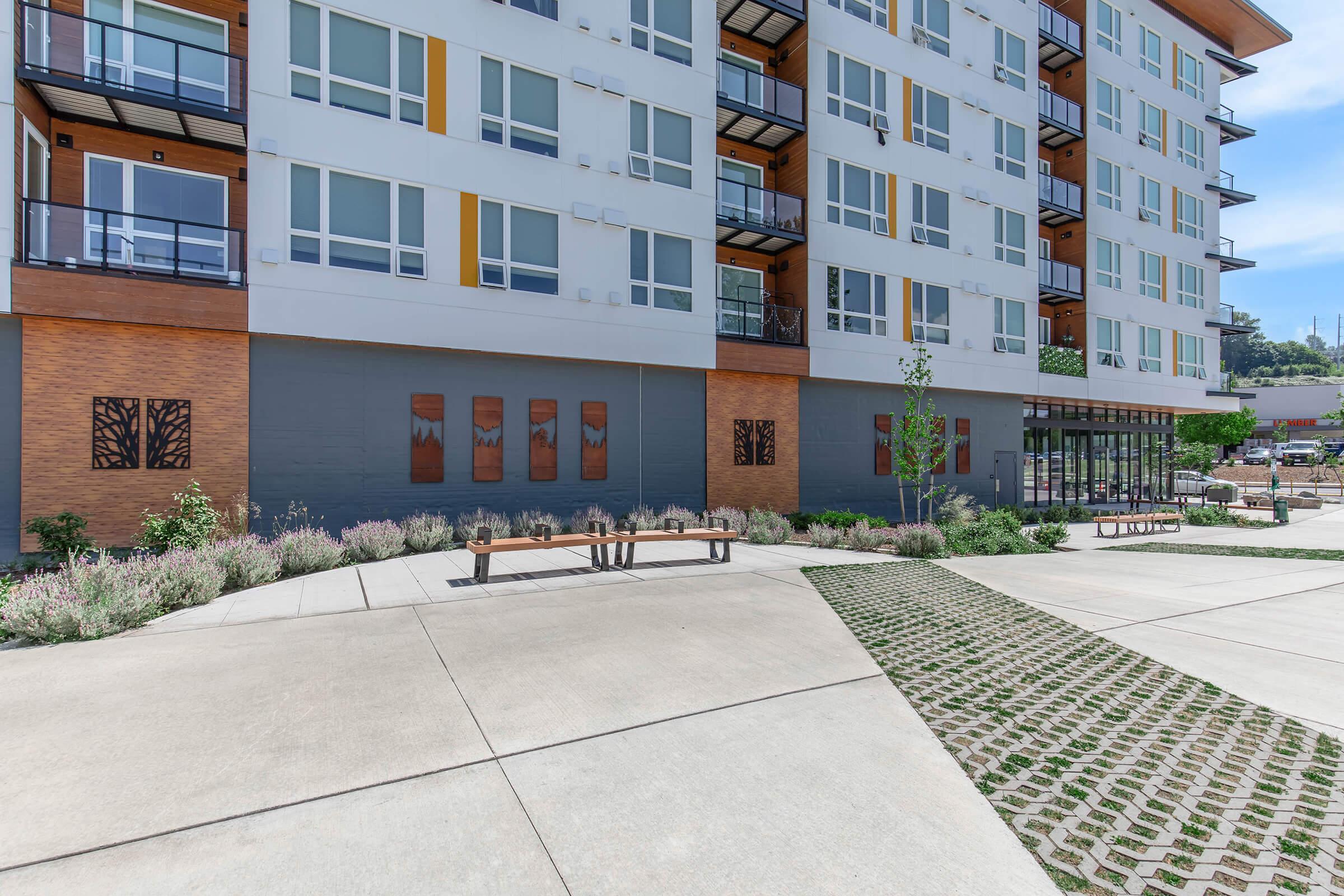 Modern apartment building with large windows and balconies, featuring a landscaped area with benches, low greenery, and artistic wall panels. The pavement consists of concrete and stone patterns, creating an inviting outdoor space. Blue sky and nearby structures are visible in the background.