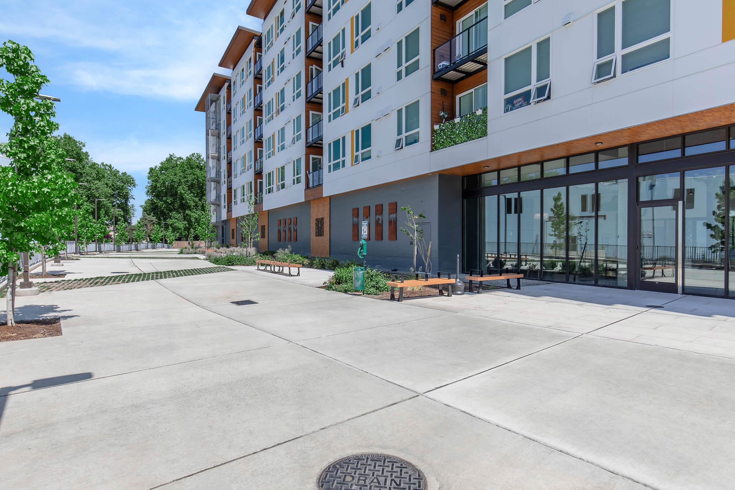A modern apartment building with multiple floors and large windows. The scene features a wide paved walkway lined with trees and benches. There are landscaped areas and seating arrangements, creating a welcoming outdoor space. Bright blue skies provide a vibrant backdrop to the contemporary architecture.