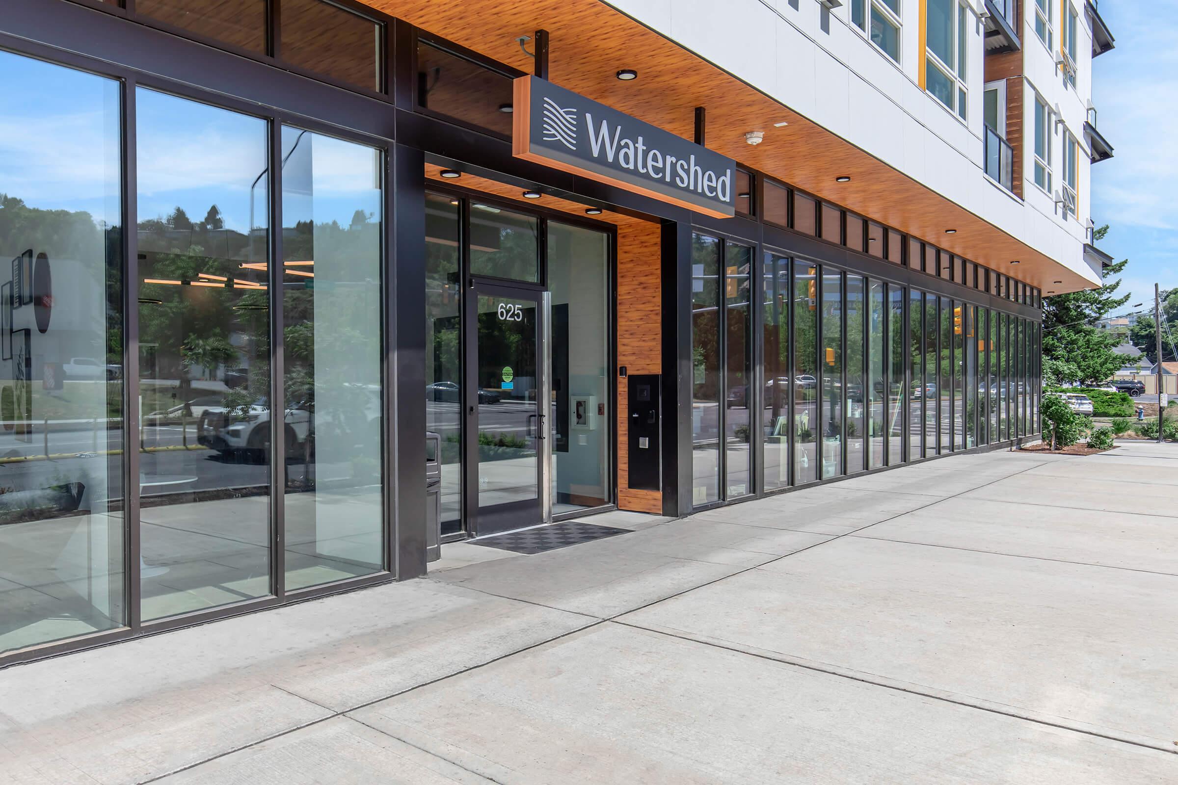 Building entrance with large glass windows and a modern design. The sign above the door reads "Watershed." The entrance is at street level with a concrete walkway, surrounded by greenery and a view of a road in the background. Bright daylight enhances the inviting atmosphere.
