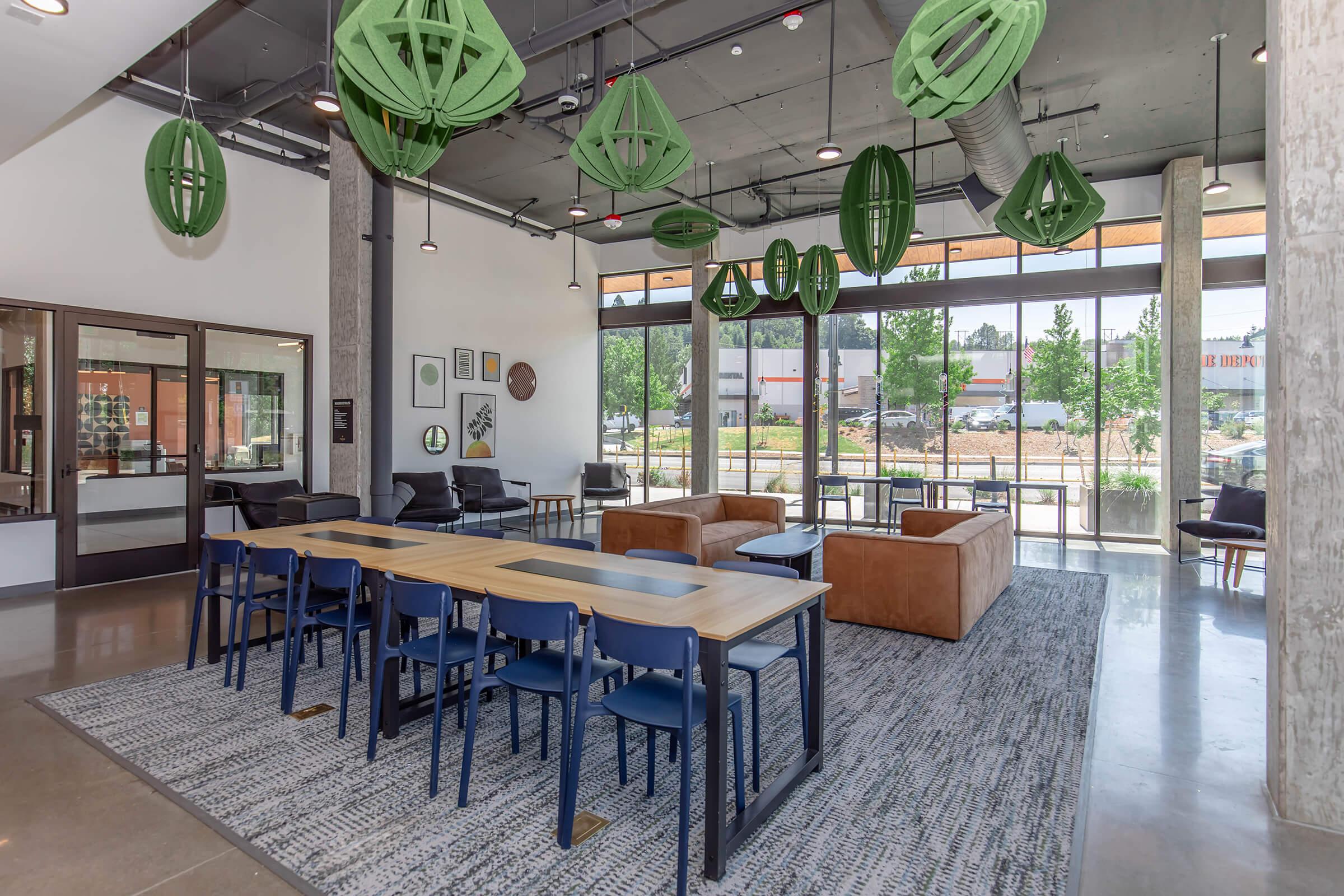 A modern lounge area featuring a long wooden table surrounded by blue chairs, plush brown sofas, and green decorative pendant lights. Large windows provide ample natural light, showcasing a view of outside greenery and parking. The space has a contemporary design with a polished concrete floor and stylish decor.