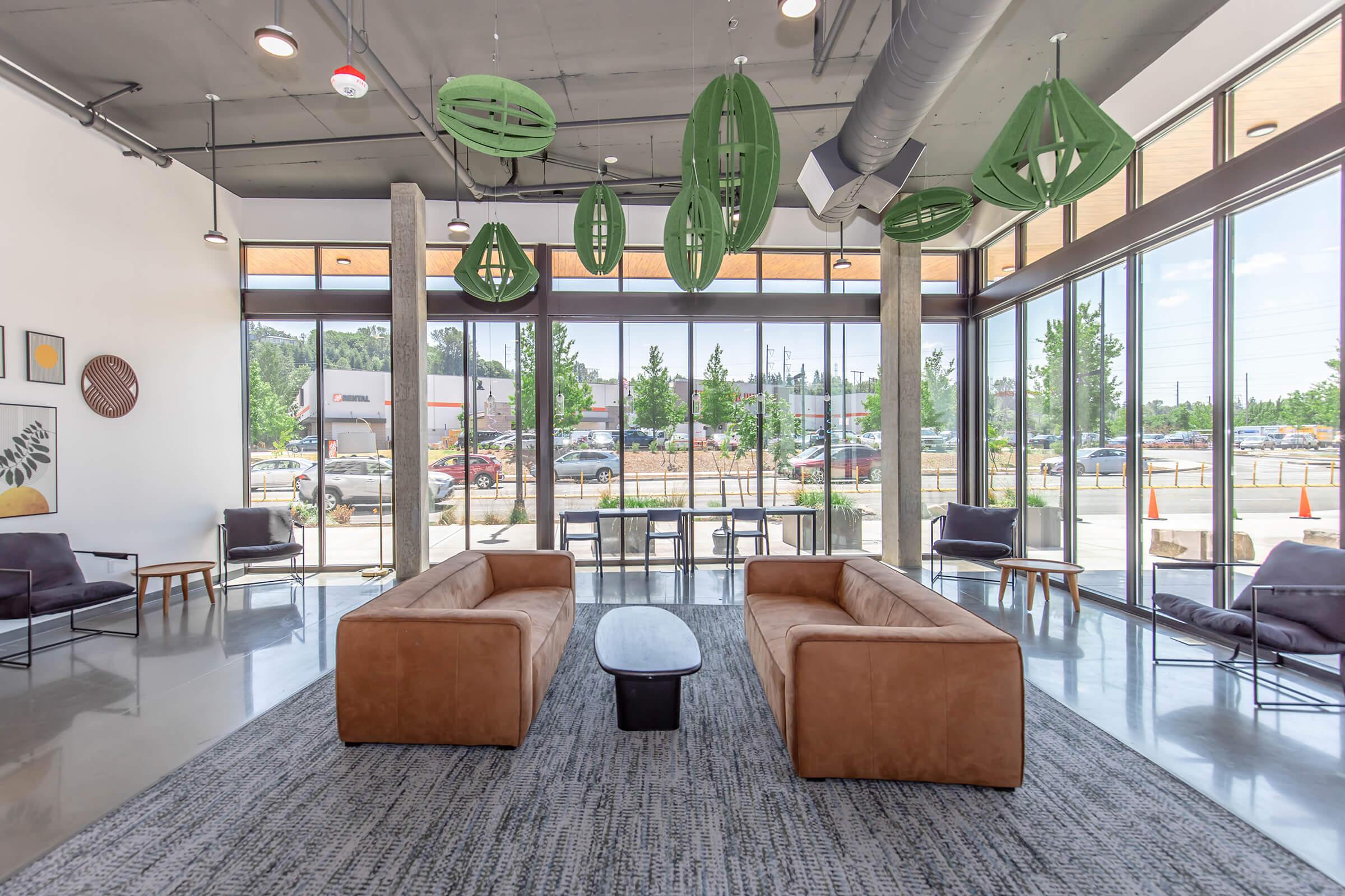 Modern lounge interior featuring two brown sofas, a black coffee table, and contemporary decor. The space is bright with large windows allowing natural light, framed by green pendant lights. Grey carpet enhances the aesthetic, while a tree-lined view is visible through the glass.