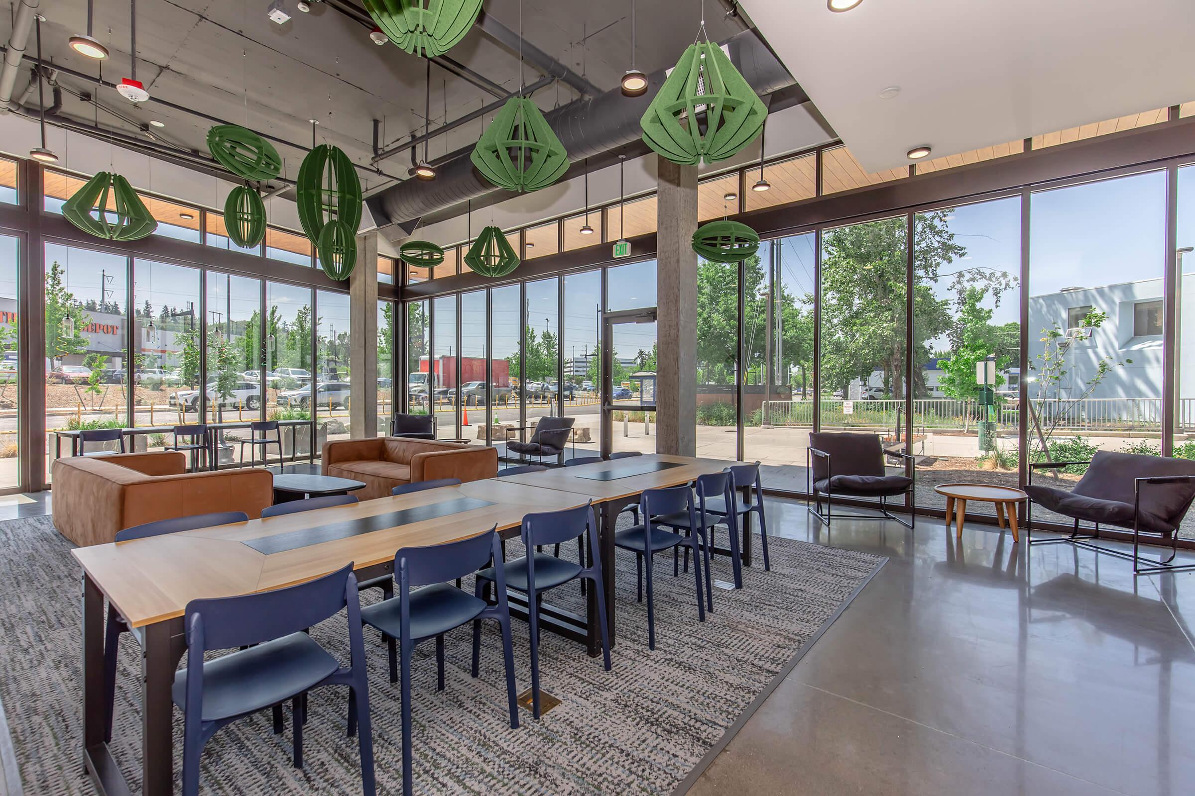 Modern indoor lounge area featuring large windows, pendant lighting with green geometric designs, and various seating options including brown couches and blue chairs. A long table is set for group use, with a stylish rug underfoot and greenery visible outside, creating a bright and inviting atmosphere.
