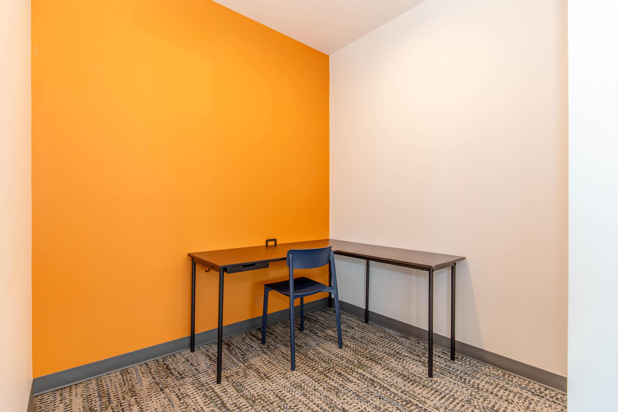 A small, minimalist room with an orange accent wall. There is a wooden desk and a single blue chair, set against the wall. The floor has a textured carpet, and the overall ambiance is simple and uncluttered.
