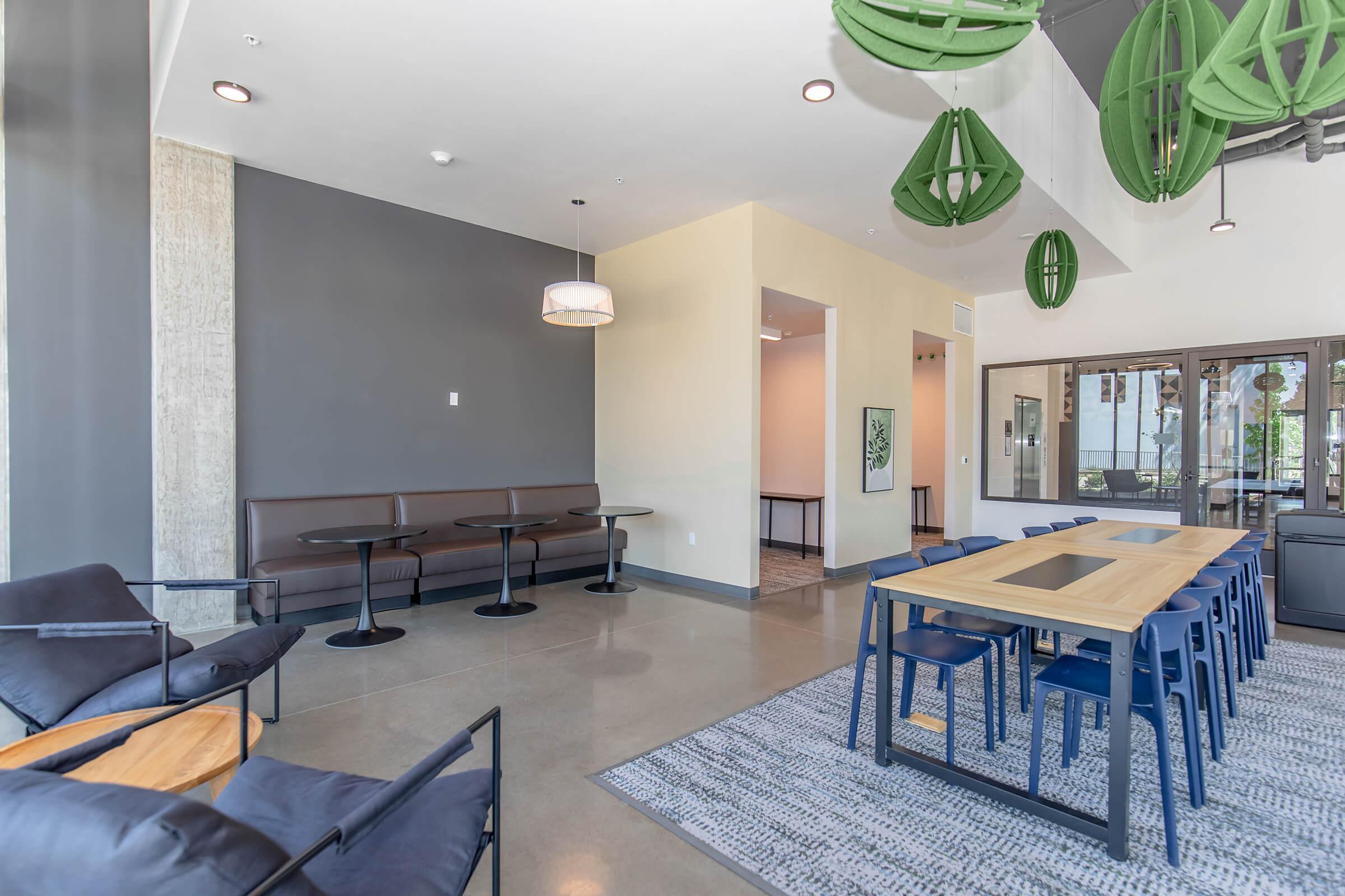 A modern communal space featuring sleek furniture, including a table with chairs, a cozy seating area with black armchairs, and green decorative hanging plants. The walls are a combination of gray and light colors, with large windows bringing in natural light. A stylish rug adds warmth to the concrete flooring.