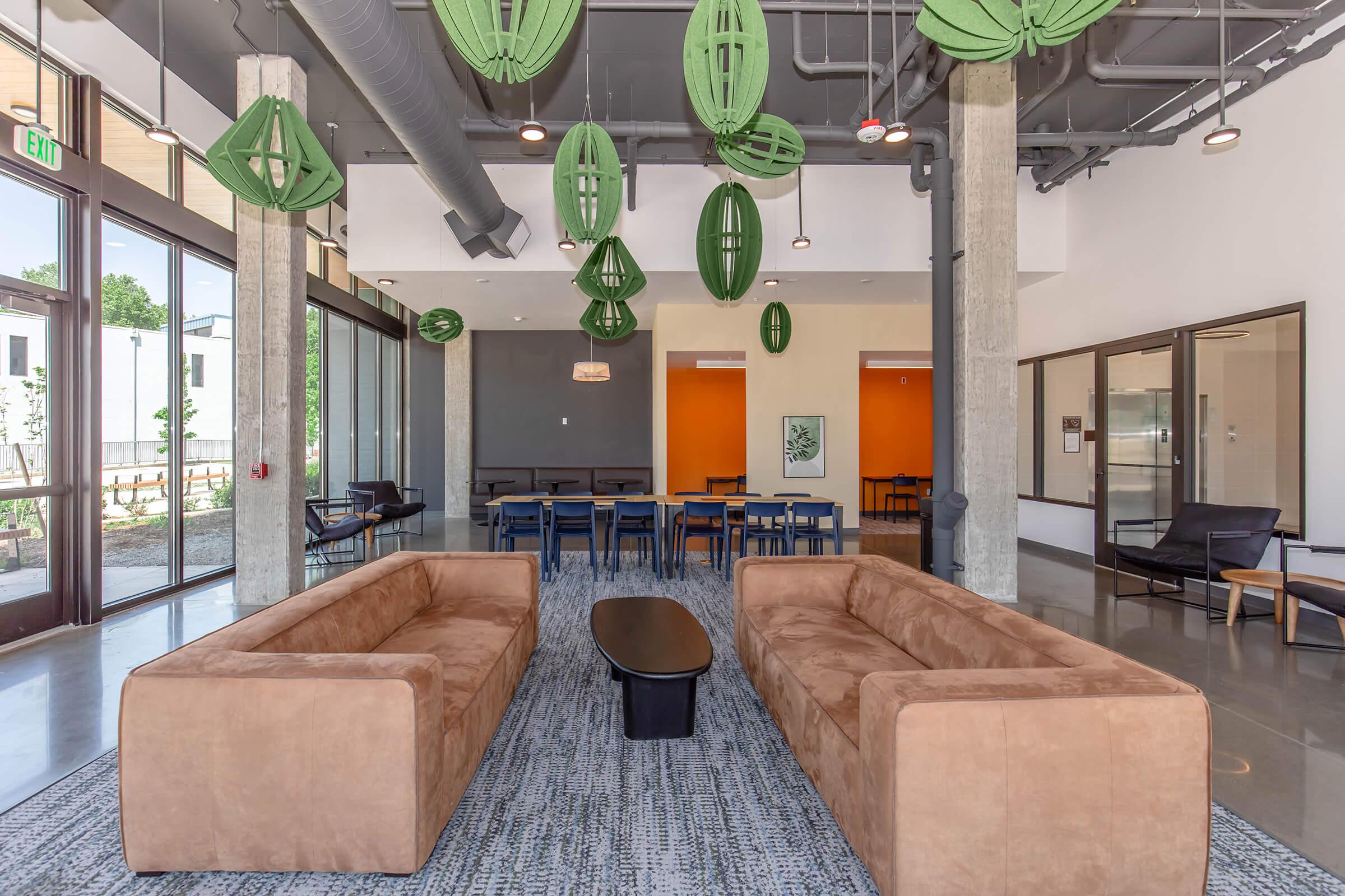 Modern interior of a lounge with two brown sofas facing a dark coffee table. The space features green hanging decorations and has large windows allowing natural light. A dining area is visible in the background with minimalist furniture, characterized by orange accent walls and an open feel.