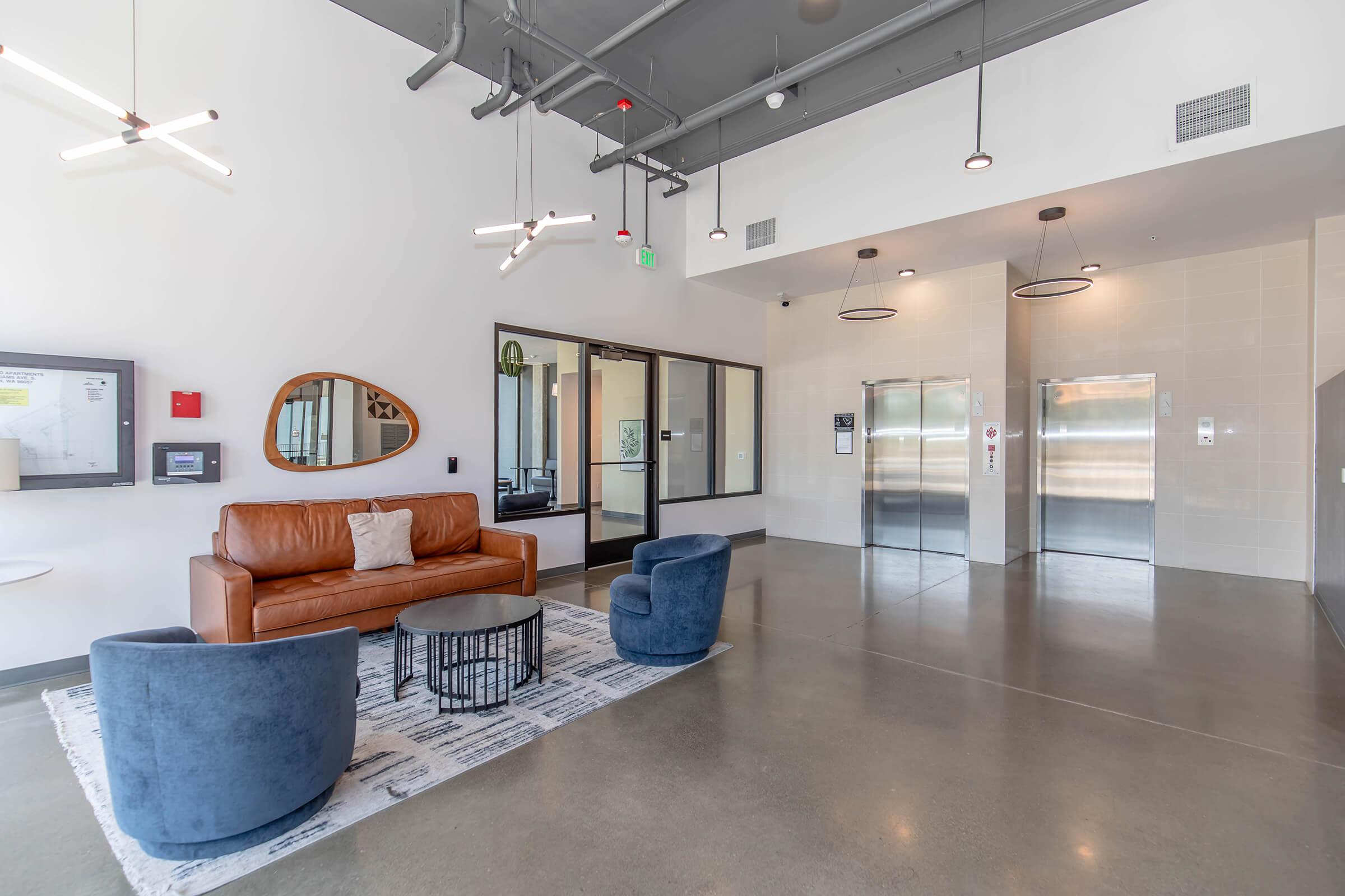 Modern lobby area featuring a brown leather sofa, two blue upholstered chairs, a black coffee table, and a rug. Large windows provide natural light, and there are two elevators in the background. Stylish lighting fixtures hang from the ceiling, contributing to a contemporary aesthetic.