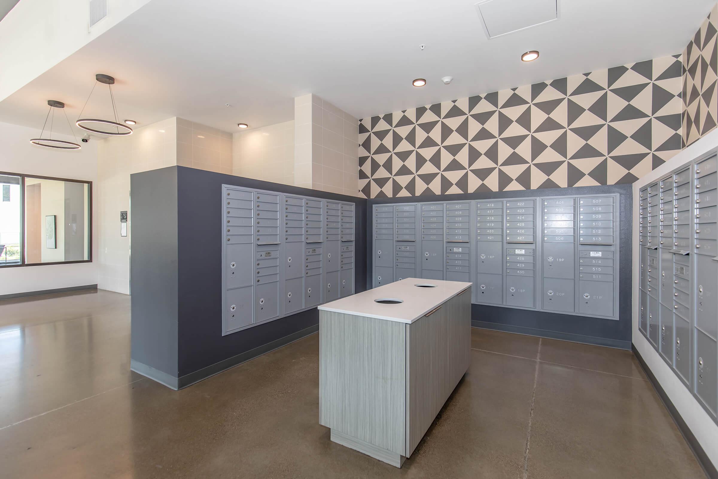 A modern mailroom featuring grey and silver mailboxes arranged in rows on a wall. In the center, there is a sleek white table with two circular openings. The walls have a decorative geometric pattern in black and white, and the floor is polished concrete. Soft lighting from pendant fixtures enhances the contemporary design.