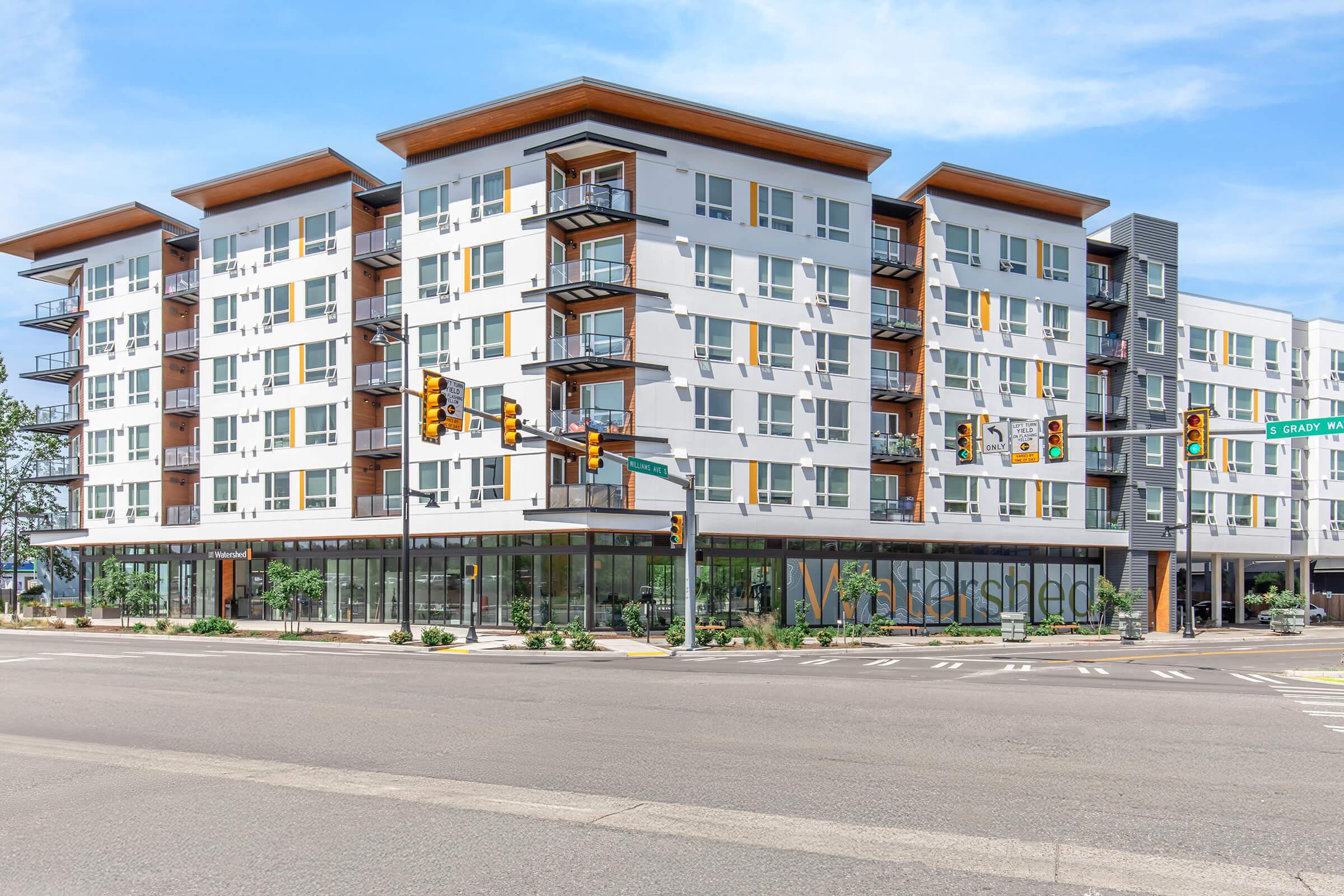 A modern multi-story apartment building with a clean, white facade featuring large windows and wooden accents. The ground floor includes a glass storefront, and the corner of the building has a sign that reads "Watershed." The scene is set at a street intersection with traffic lights.