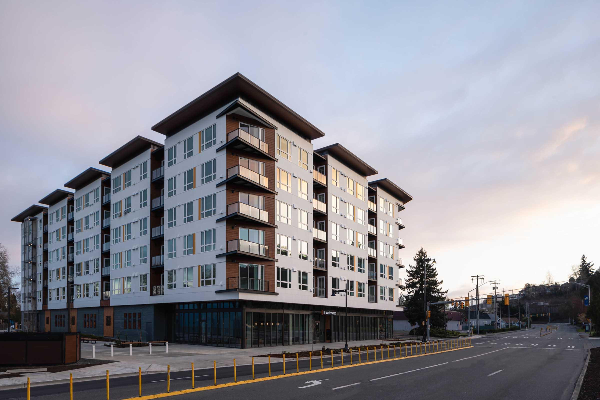 Modern multi-story apartment building with large windows and balconies, situated on a wide street. The structure features a contemporary design with a mix of light and dark exterior materials. The sky above is cloudy and hints at early evening light.