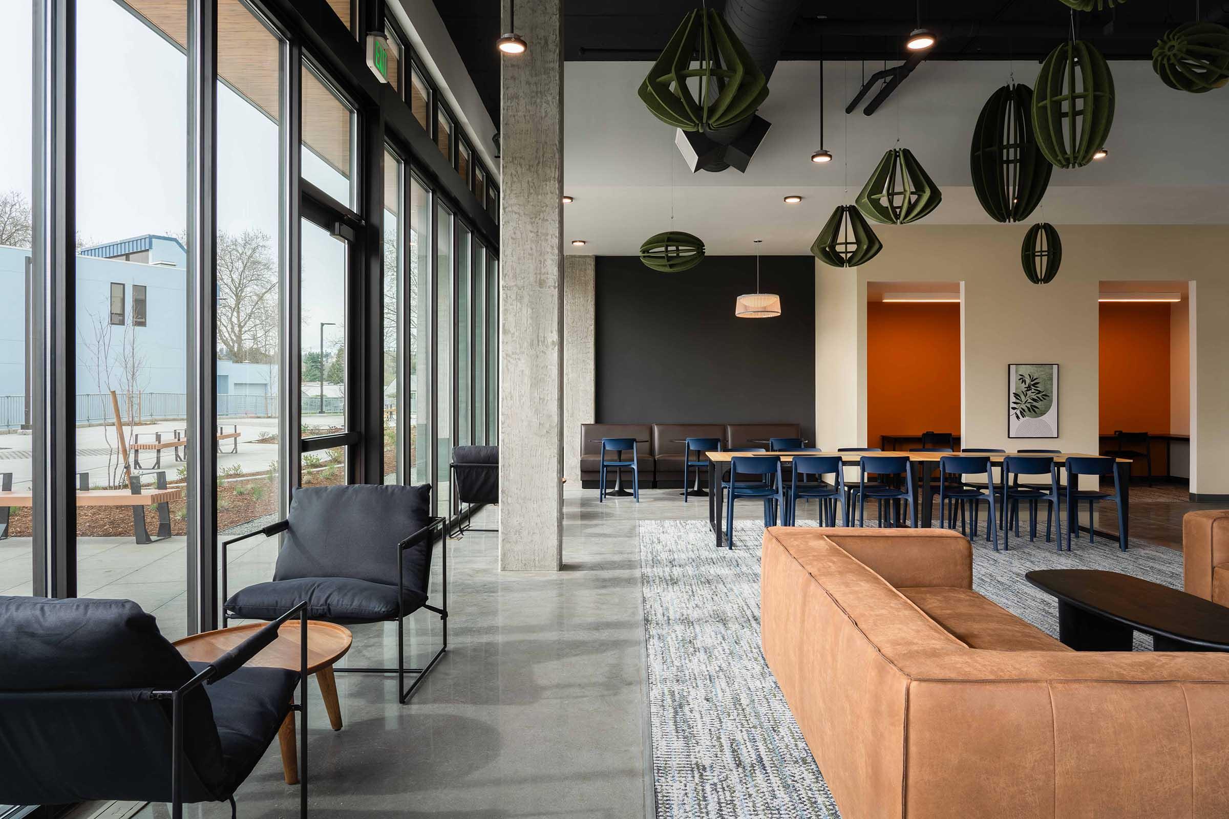 Modern indoor lounge area featuring large windows, a mix of green and black pendant lights, and contemporary furniture. The space includes a cozy brown sofa, black armchairs, a wooden side table, and several dining tables with blue chairs. Light-colored walls and polished concrete flooring enhance the inviting atmosphere.