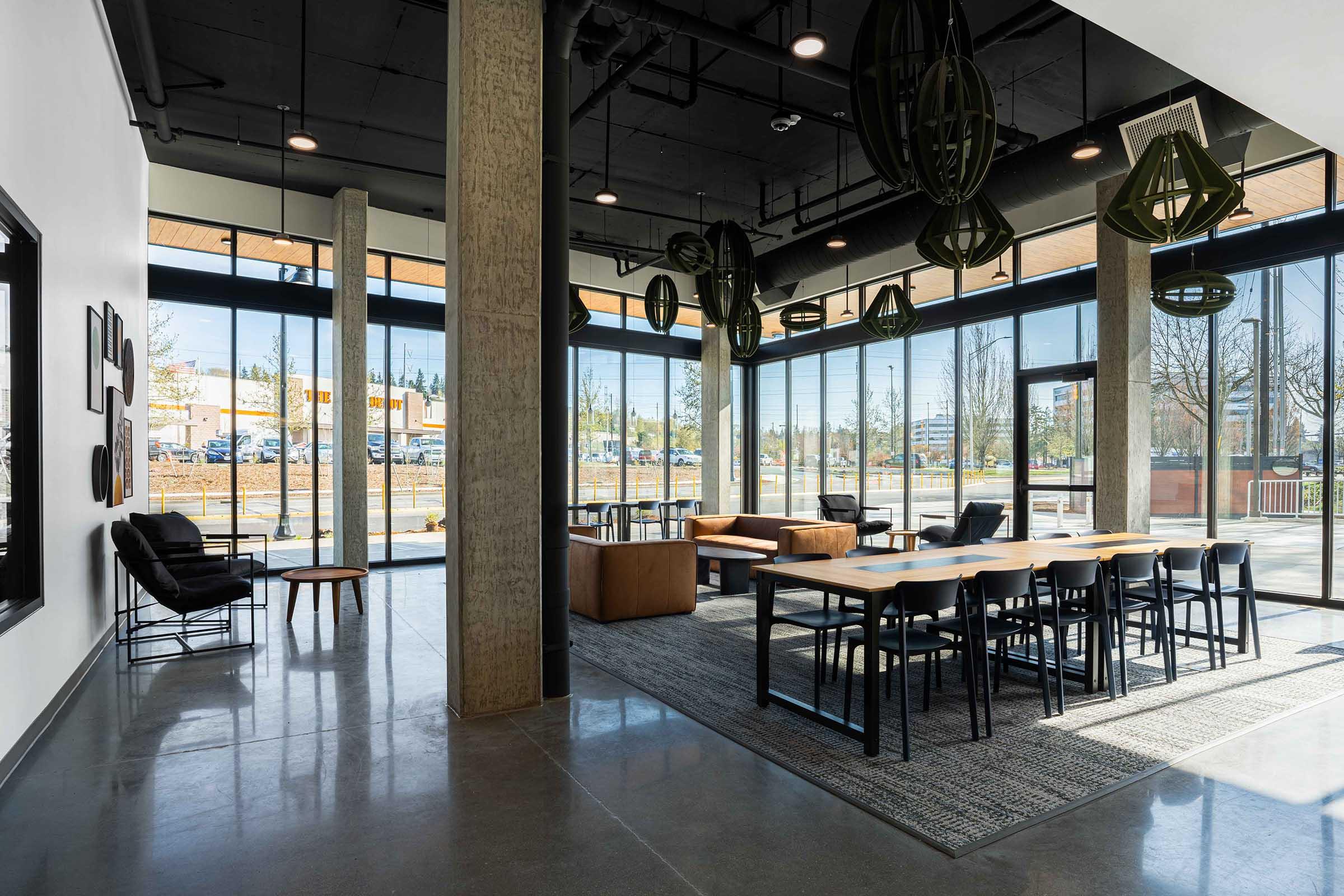 A modern interior space featuring large windows, natural light, and a mix of seating options. There are wooden tables and chairs, a cozy seating area with black chairs, and stylish light fixtures hanging from the ceiling. The floor is polished concrete, enhancing the contemporary feel of the room.