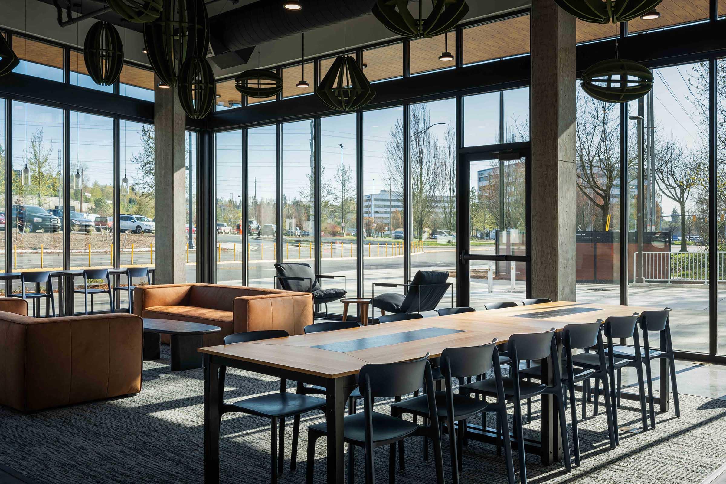 A modern indoor space featuring large windows that let in plenty of natural light. There's a wooden dining table surrounded by black chairs, and a cozy seating area with brown leather couches. Decorative pendant lights hang from the ceiling, creating an inviting atmosphere.