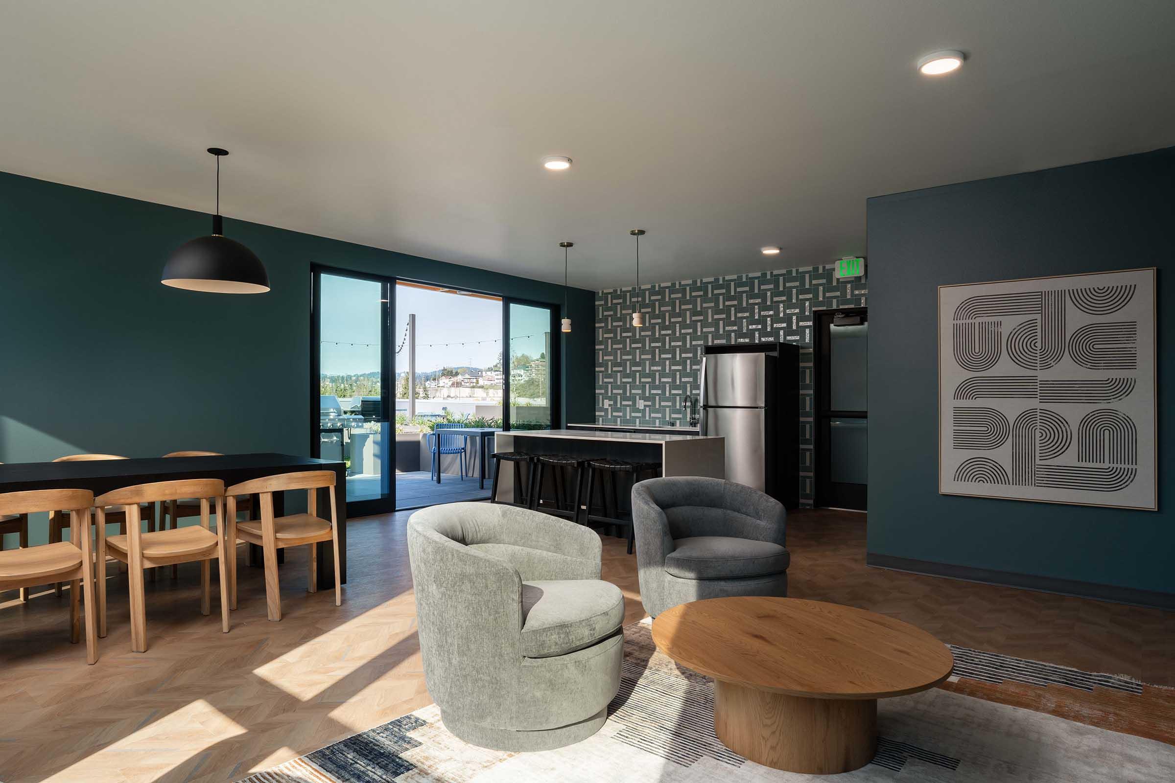 A modern living space featuring a dining area with a black table and light wooden chairs, two cozy gray armchairs, and a round wooden coffee table. The walls are painted in a deep teal color, and there's a patterned accent wall. Large windows provide natural light and outdoor views, along with a sleek kitchen area.