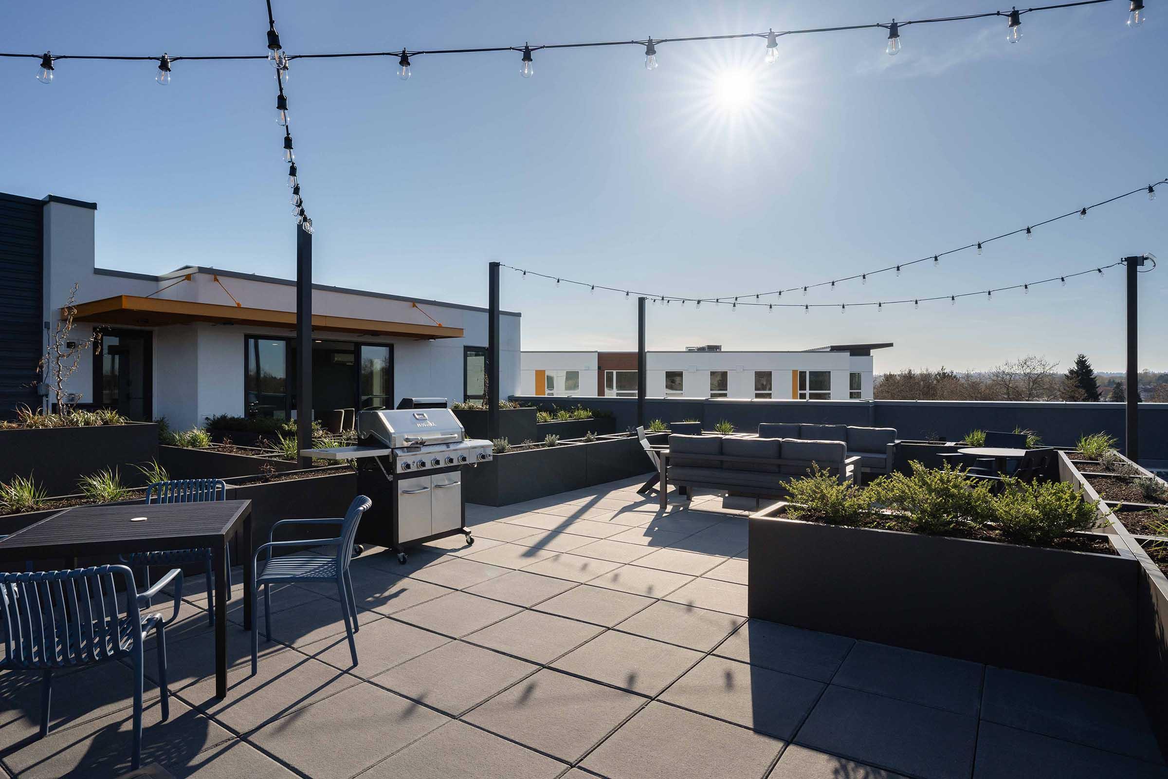 Rooftop terrace featuring patio seating, a barbecue grill, and potted plants, illuminated by sunlight with string lights overhead. The scene includes modern architectural elements and a clear blue sky in the background.