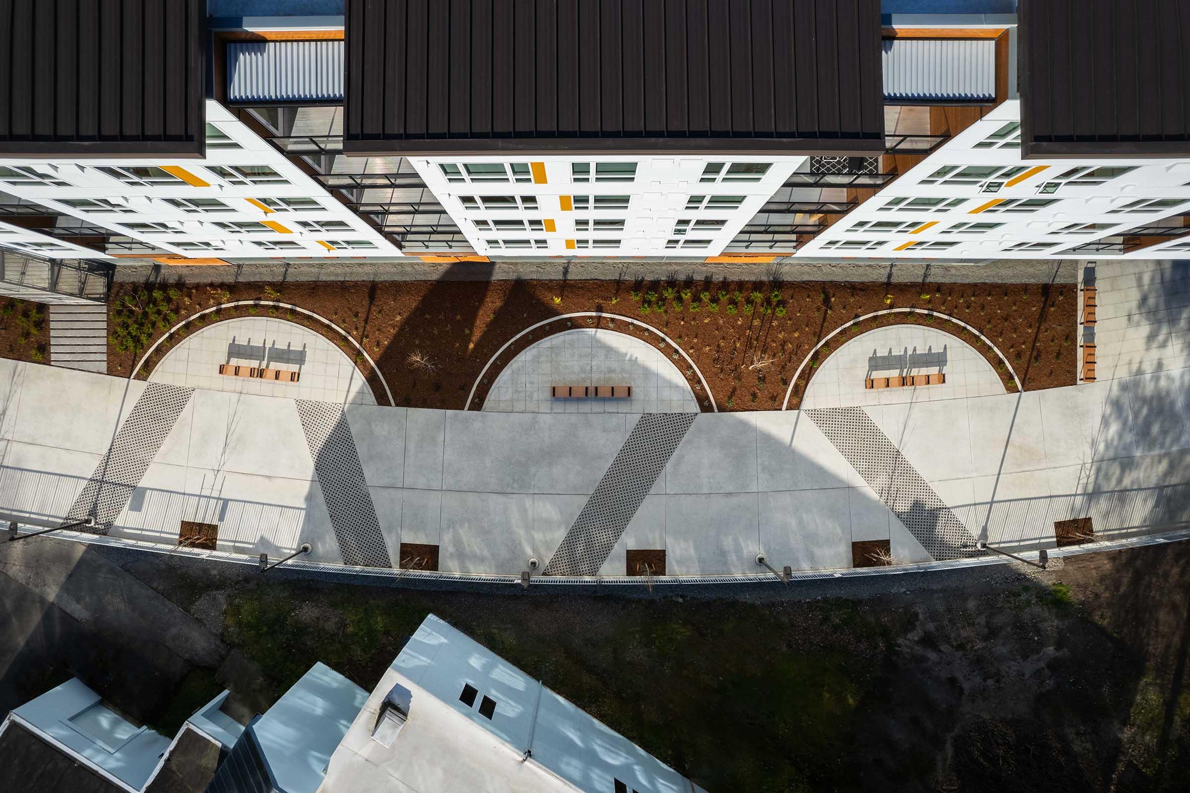 Aerial view of a modern building with a landscaped courtyard below. The courtyard features circular garden beds, benches, and paved walkways, intersecting with shadows cast by the building above. Surrounding greenery is visible, enhancing the outdoor space's inviting atmosphere.