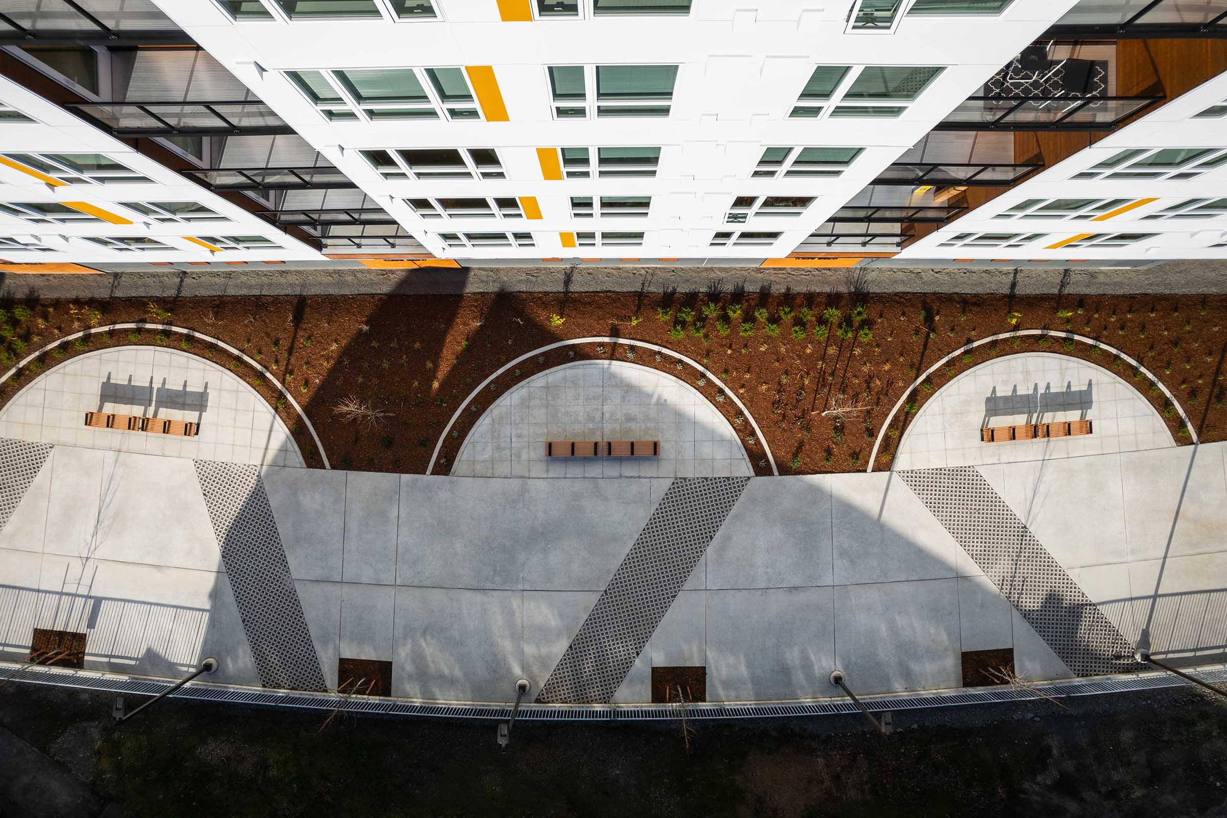 Aerial view of a modern building with white facade and orange accents, showcasing landscaped circular seating areas on the ground below. The pavement includes curved paths and benches amidst greenery, creating an inviting outdoor space. Sunlight casts shadows, enhancing the design's geometric patterns.