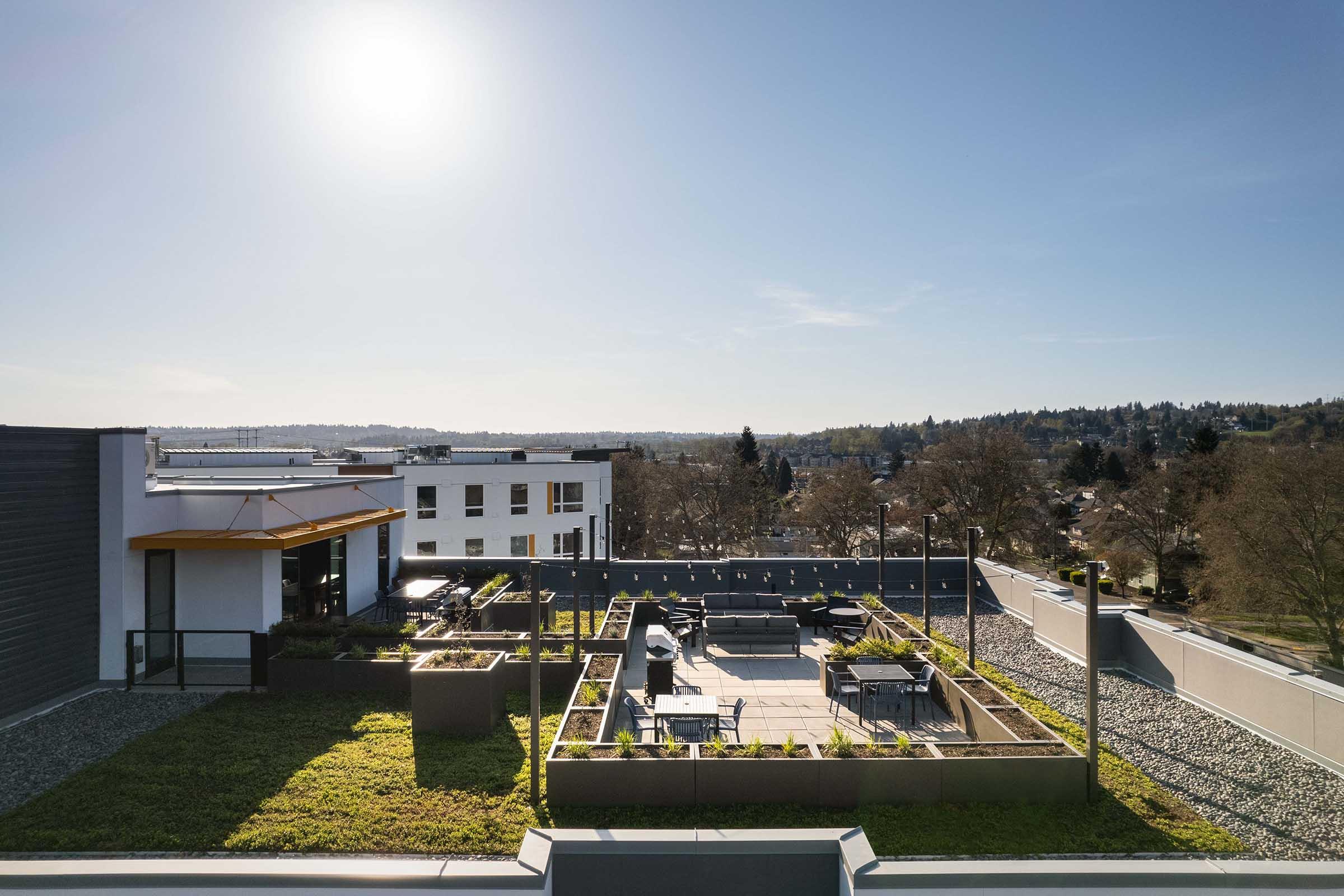 A rooftop terrace featuring landscaped gardens, seating areas, and sunny views of the surrounding cityscape. The scene captures a clear blue sky with the sun shining, creating a bright and inviting atmosphere for outdoor relaxation.