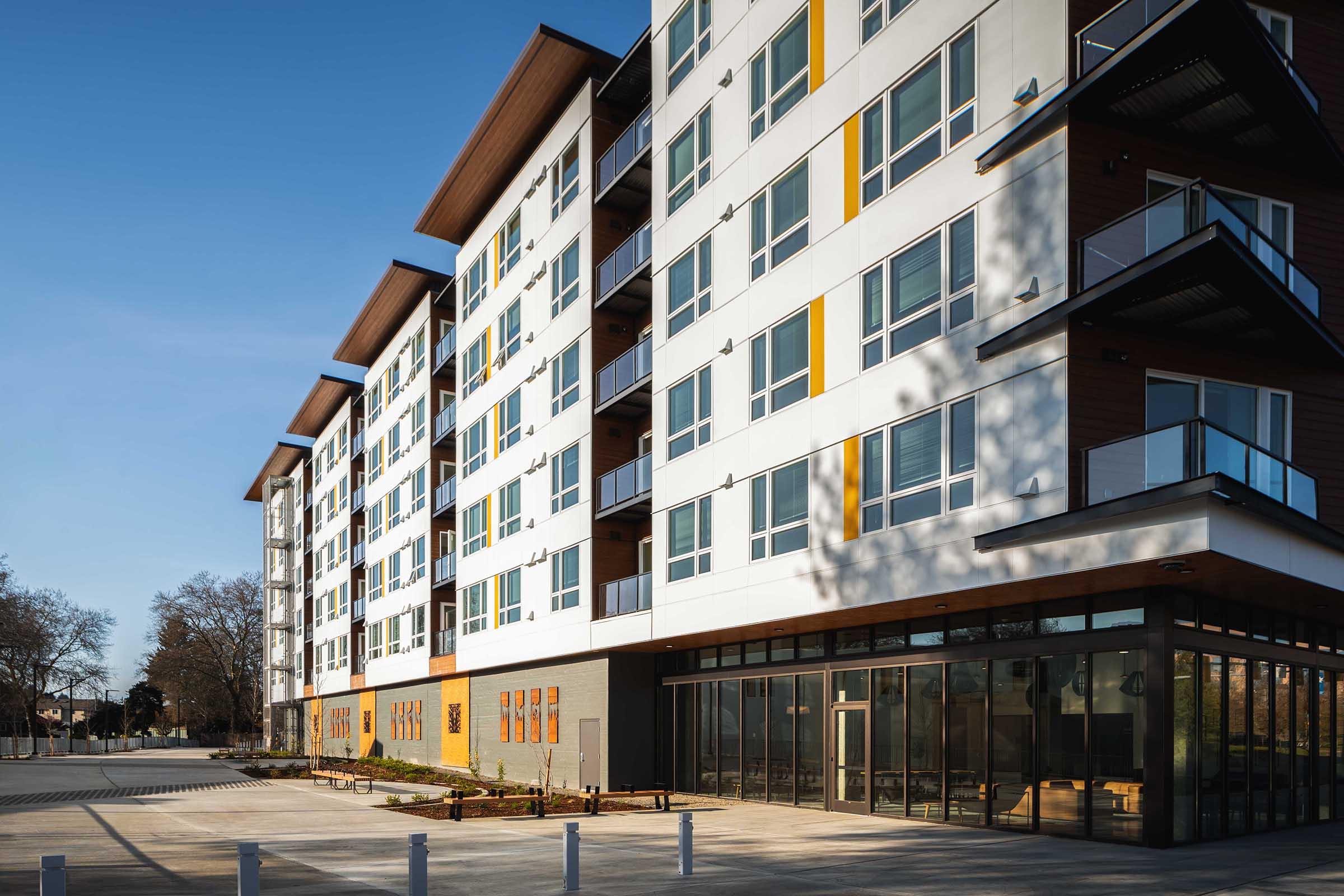 Modern multi-story building with a sleek design featuring large windows and balconies. The structure is surrounded by a paved area, with trees in the background and a clear blue sky. The first floor includes expansive glass windows indicating communal or retail space.