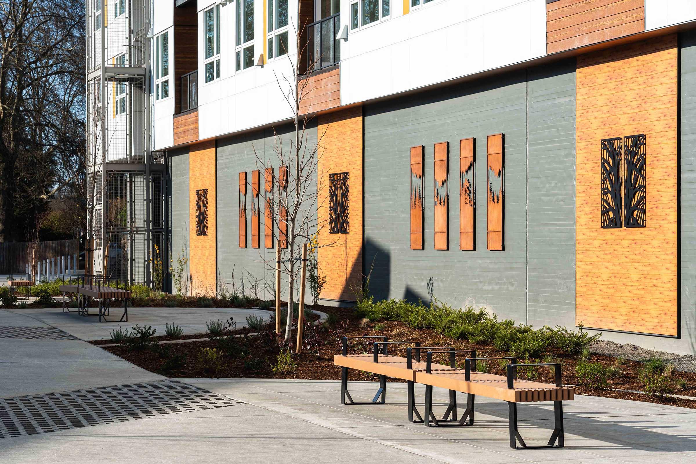 A modern building exterior featuring a landscaped area with benches and decorative wall panels. The wall has wooden and metal art installations, surrounded by greenery and a paved walkway. Natural light illuminates the scene, enhancing the contemporary design elements.