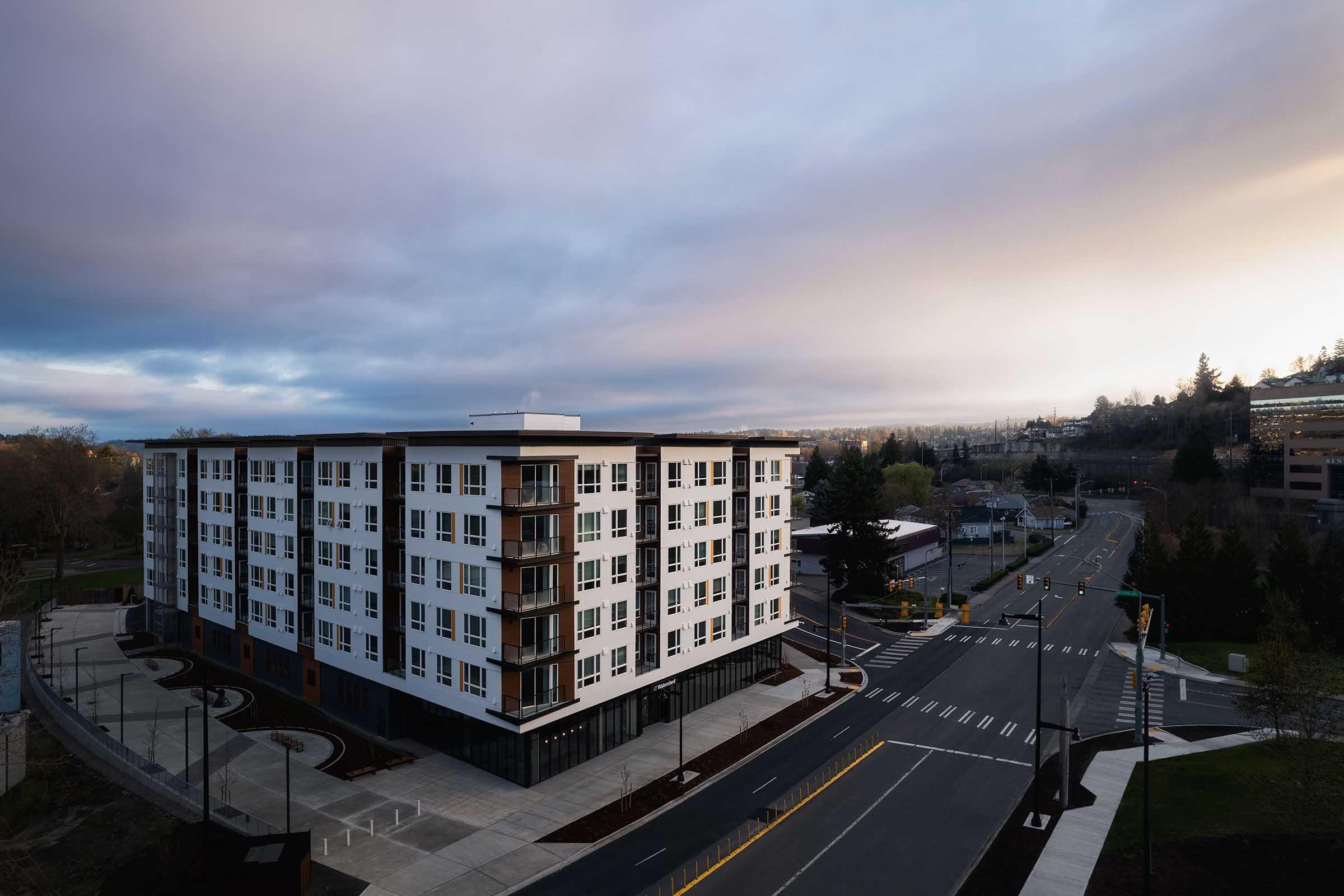 A modern multi-story apartment building with a sleek white facade. The building features balconies and large windows, situated at the corner of a street. The background shows a cloudy sky and a cityscape, with roads and greenery surrounding the area.