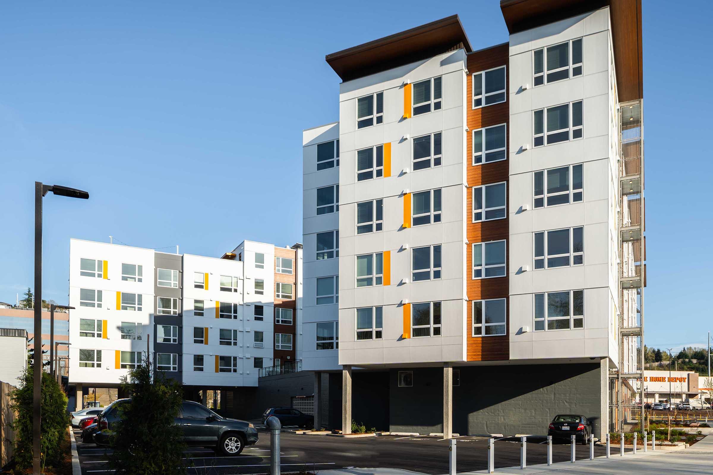 Modern multi-story residential apartment building featuring a mix of white and wooden paneling. The structure includes large windows with yellow accents and is surrounded by a paved parking area and landscaped greenery. A clear blue sky is visible in the background, emphasizing the bright and contemporary design.