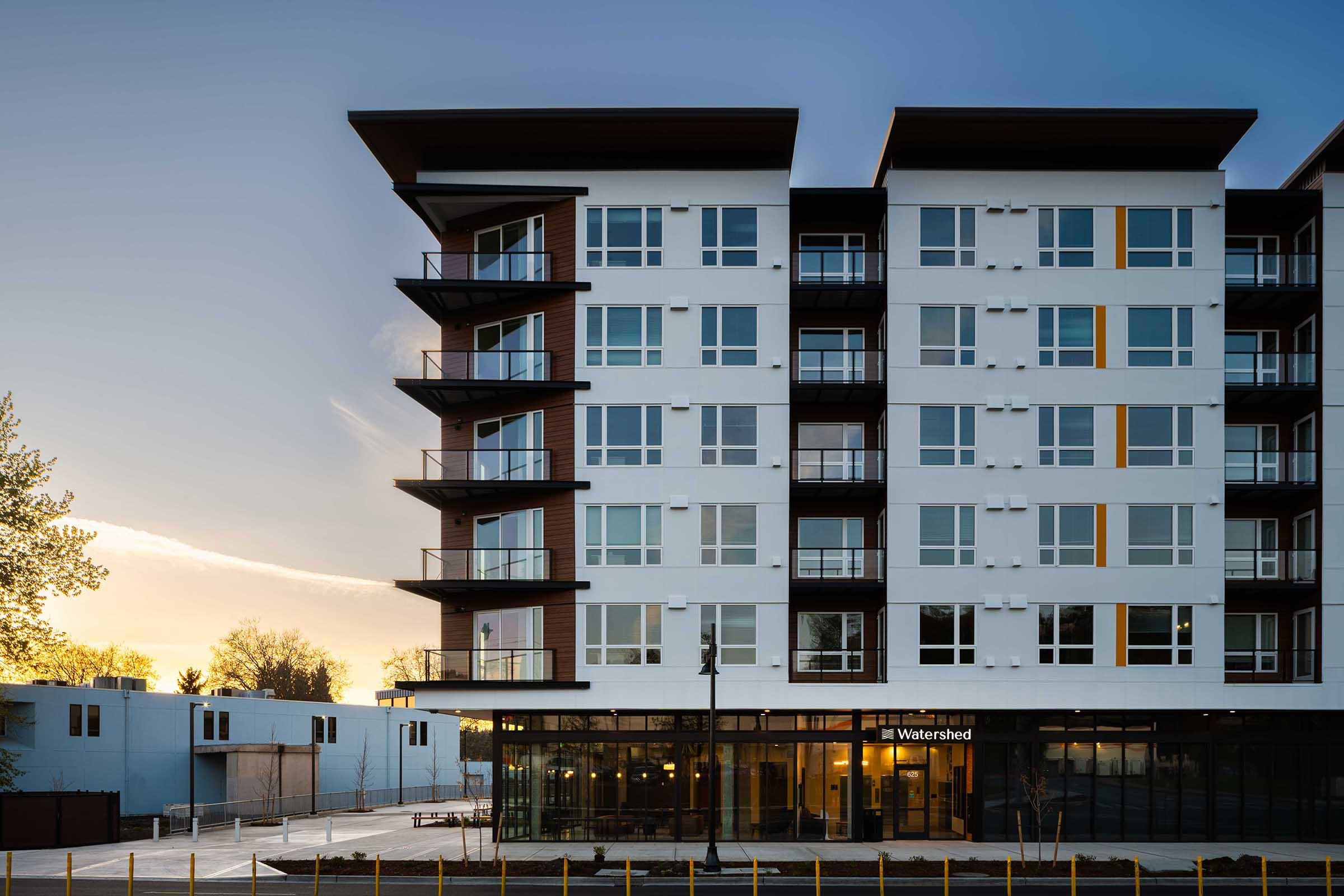 Modern residential building with a contemporary design featuring multiple balconies and large windows, set against a sunset sky. The ground floor includes commercial space with large glass windows. Surrounding area shows additional buildings and a clear atmosphere.