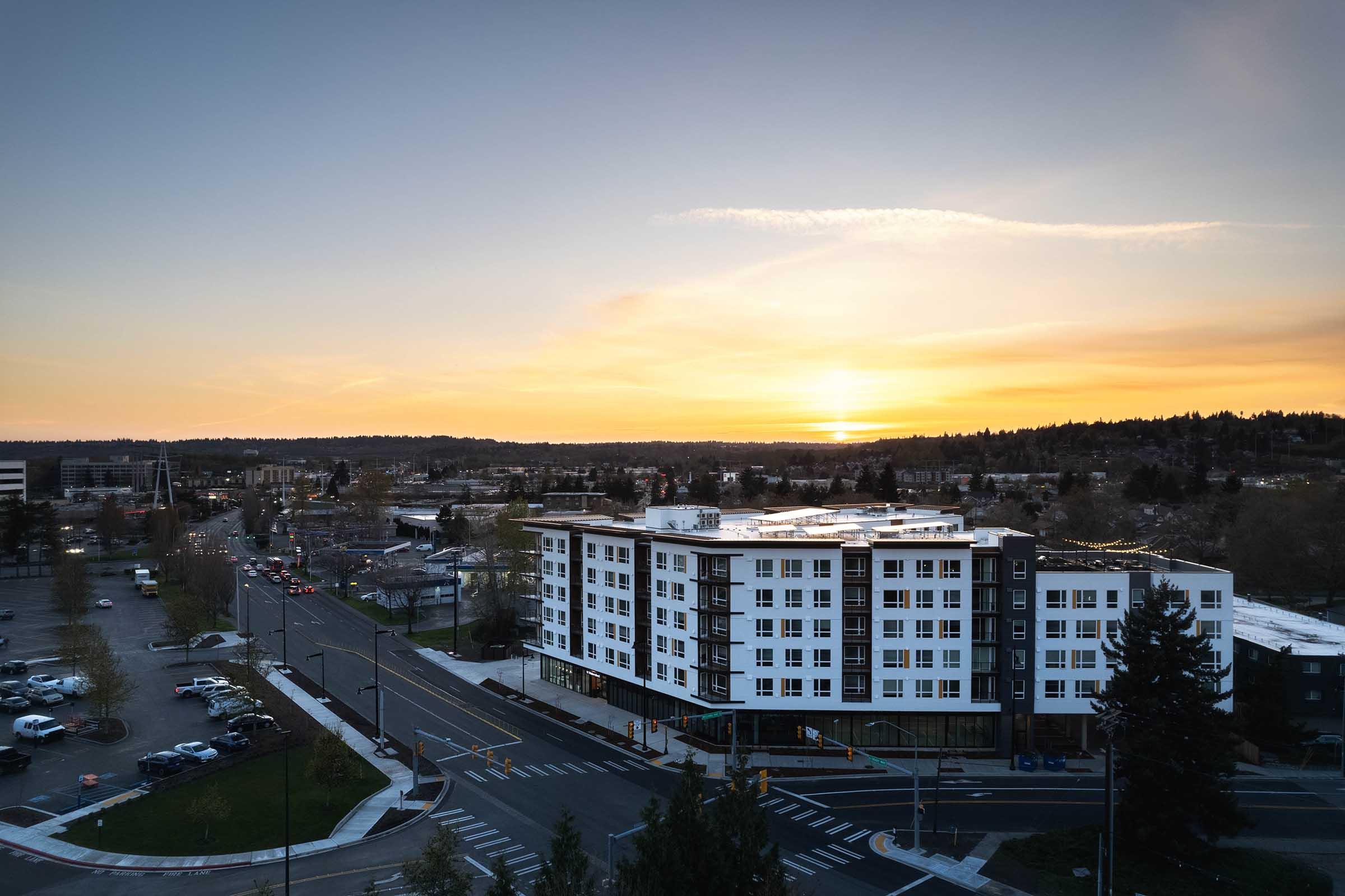 A modern multi-story apartment building near a busy intersection at sunset. The sky is filled with warm hues of orange and yellow, casting a glowing light over the landscape. Vehicles are parked nearby, and the scene features a mix of urban and natural elements with trees lining the streets.