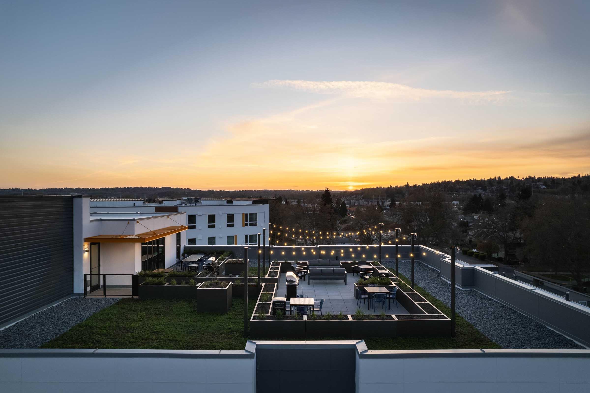 A rooftop terrace view at sunset, featuring a landscaped area with tables and seating under string lights. The horizon showcases a colorful sky as day transitions to evening. Nearby buildings provide a modern urban backdrop. The overall scene conveys a tranquil and inviting atmosphere.