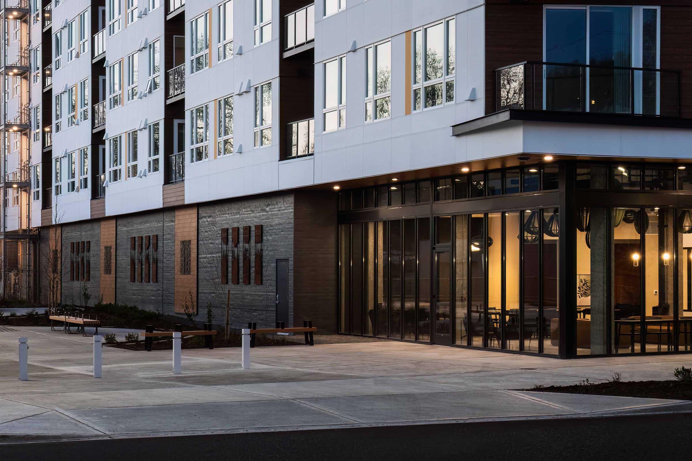 Modern apartment building exterior featuring a mix of large windows and a glass entrance on the ground floor. The pavement in front is well-defined with benches and light posts, surrounded by landscaped areas. The building has a contemporary design with contrasting materials and colors.