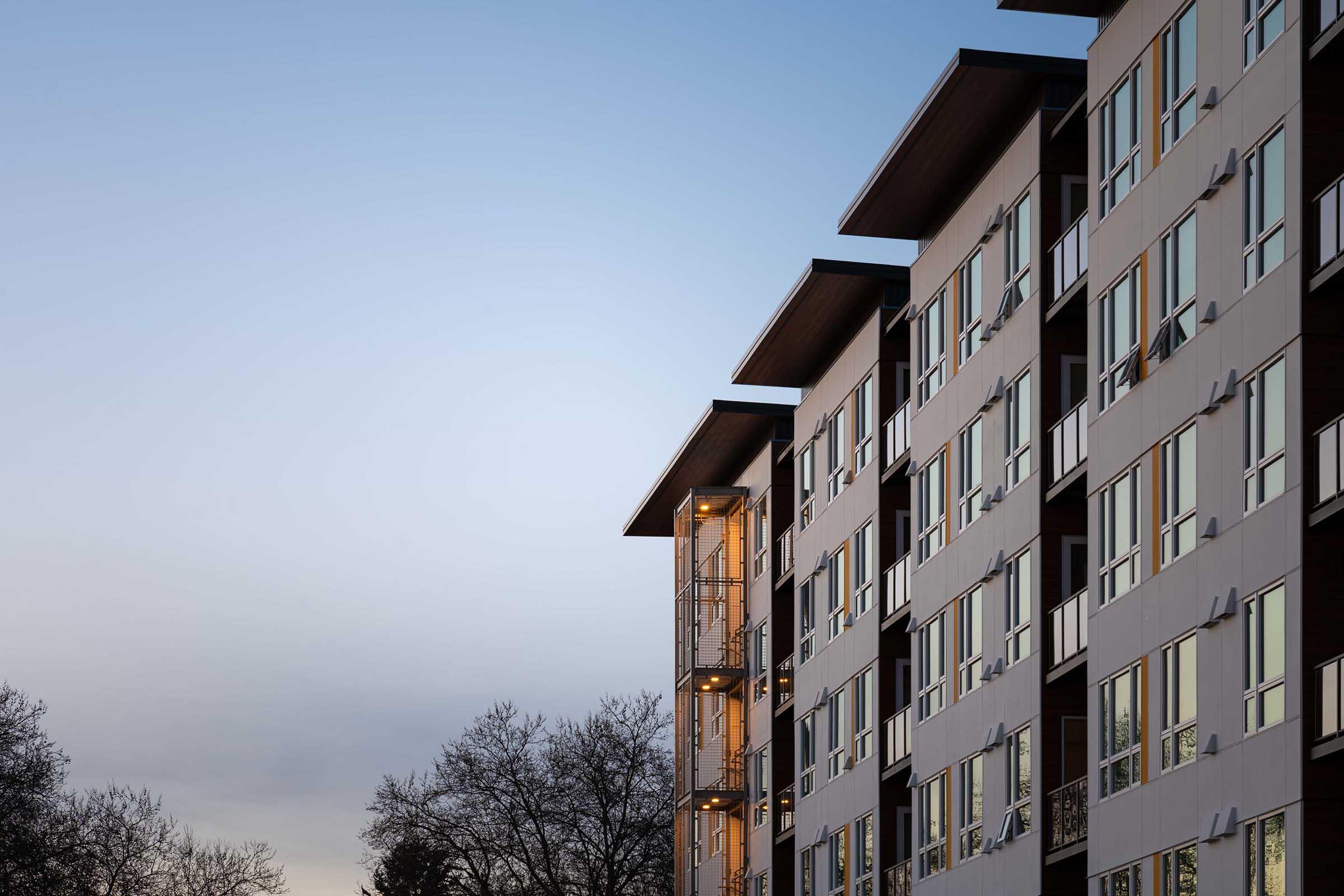 A modern apartment building featuring several floors with large windows reflecting the evening sky. The structure's design includes balconies and a mix of light and dark exterior colors. Trees are visible in the foreground, framed against the building.