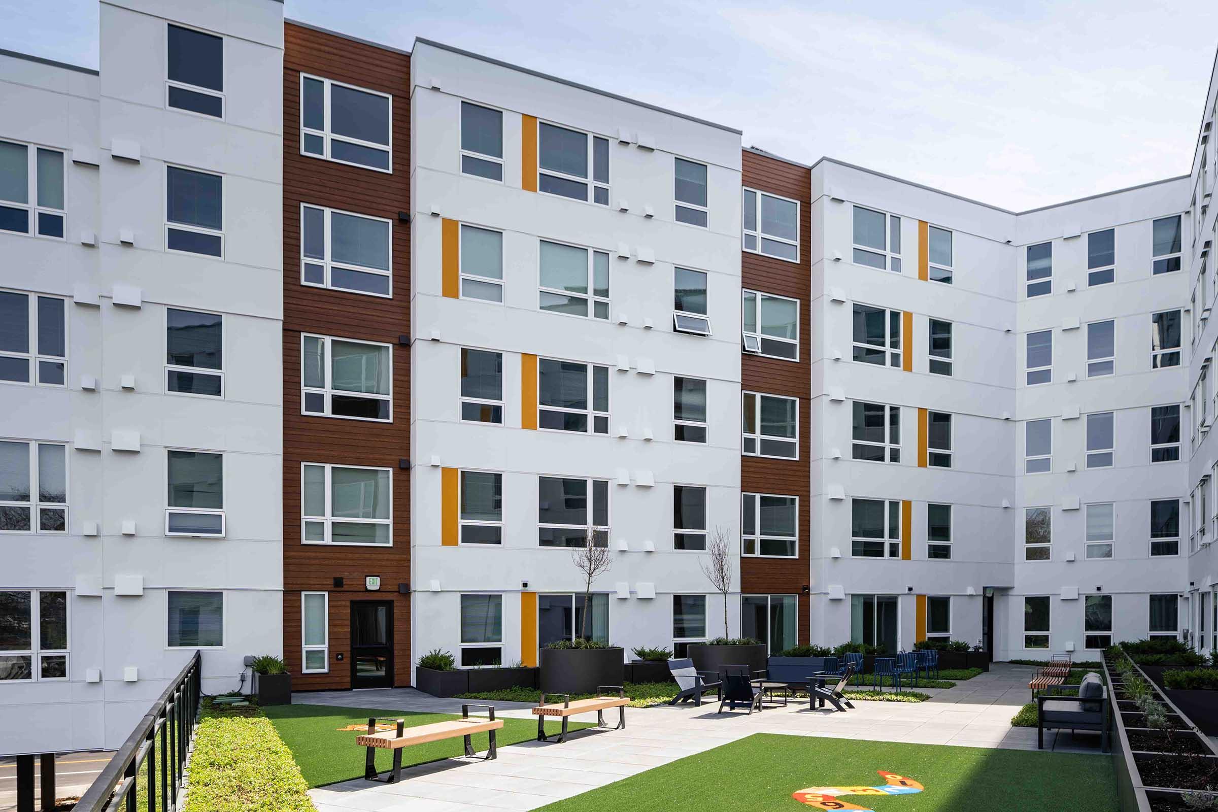 A modern apartment building with a central courtyard. The exterior features a mix of white and brown colors, with large windows and balconies. The courtyard includes seating areas and green landscaping, creating a welcoming outdoor space for residents.