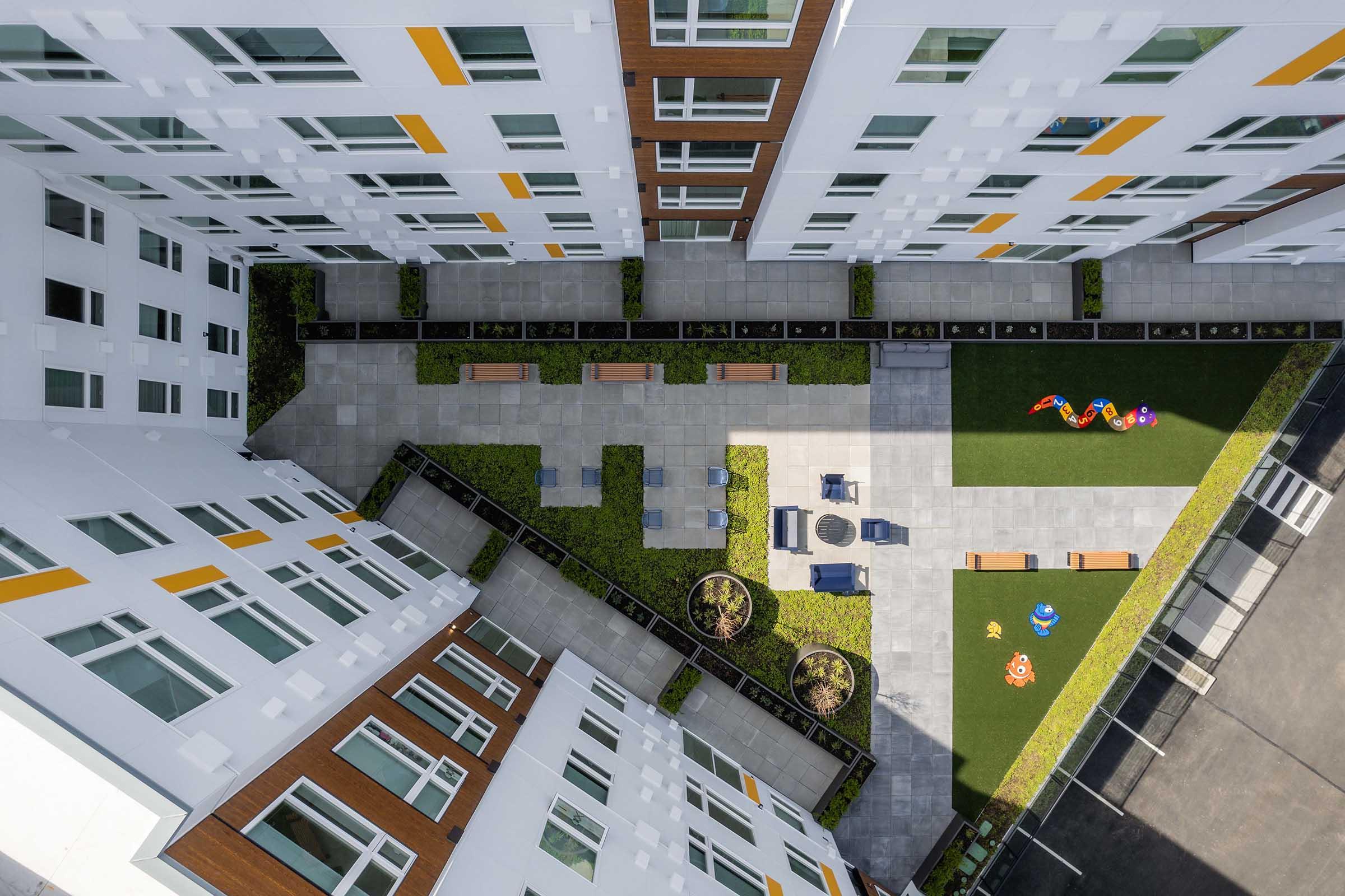 Aerial view of a modern residential building complex featuring multiple floors with large windows. The courtyard includes landscaped areas, benches, and a small playground with colorful equipment on green grass, surrounded by paved walkways and plant borders.